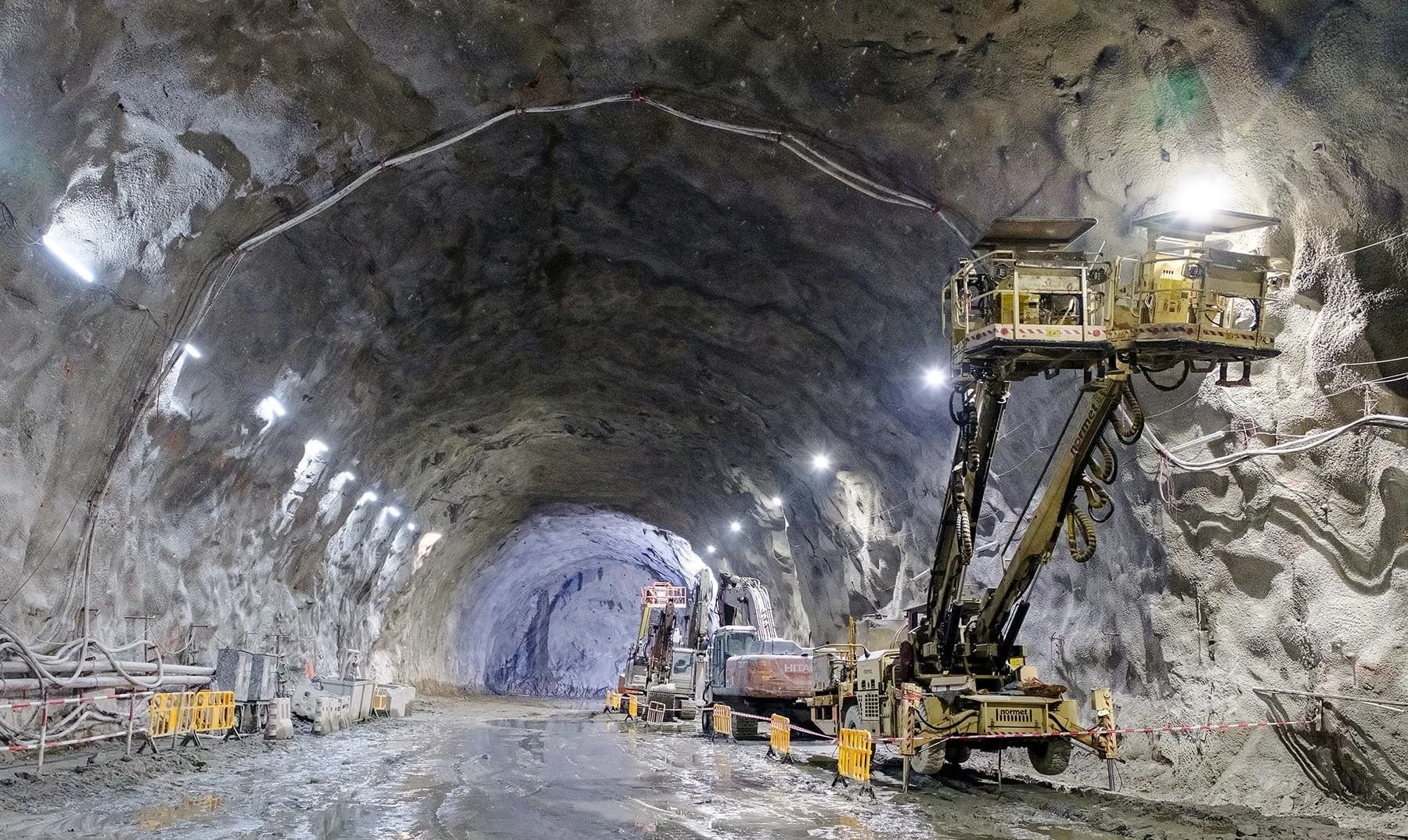 Un tunnel souterrain brut de décoffrage, éclairé par des projecteurs intenses, est rempli d'engins de chantier jaunes, dont une foreuse imposante et des excavatrices, sur un sol boueux.