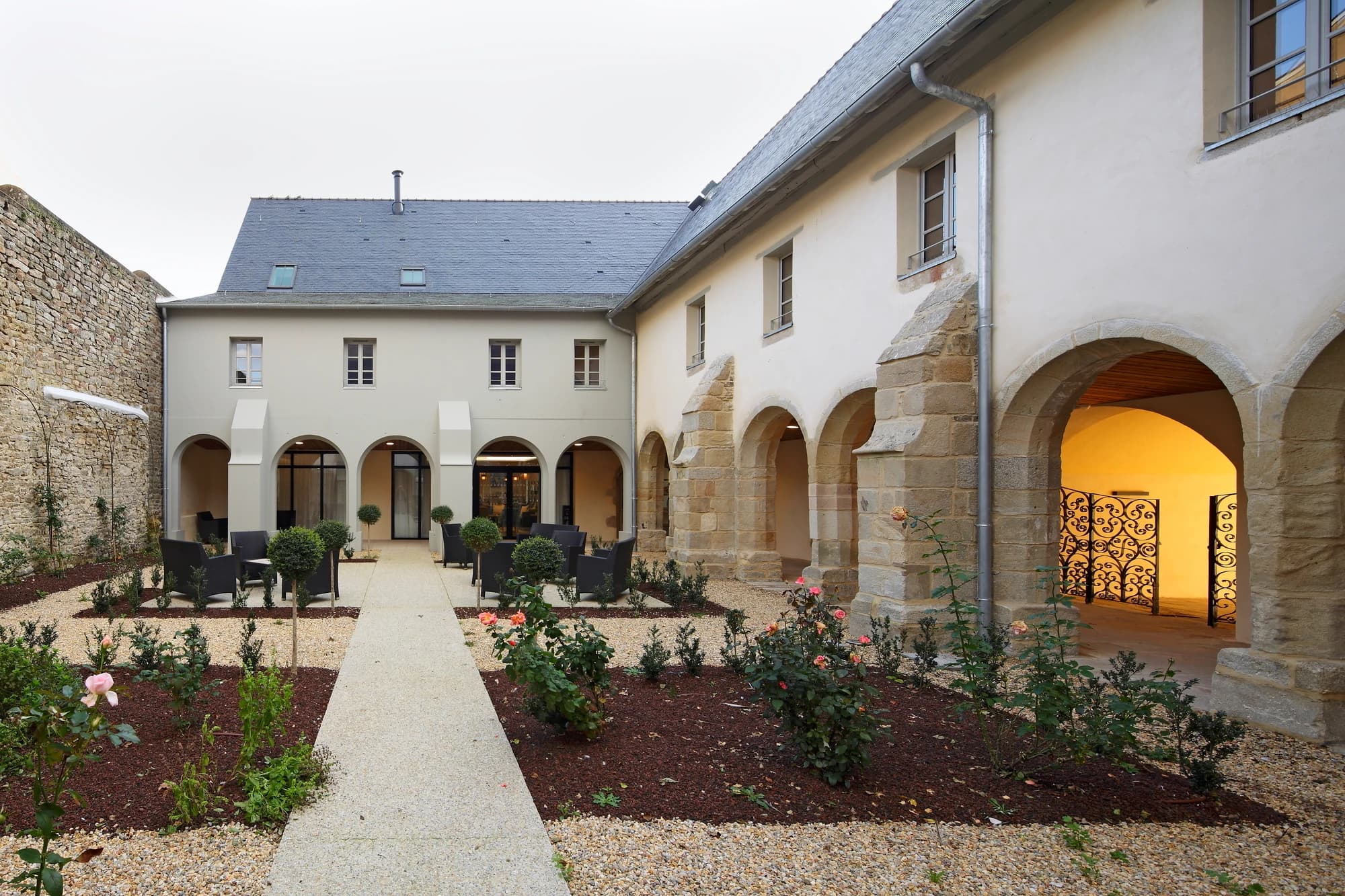 Un cloître ou une cour intérieure tranquille avec un chemin de gravier clair, des parterres de fleurs paillés agrémentés de rosiers roses et des sièges modernes sombres. Le tout est entouré de bâtiments historiques aux murs crème pâle, aux toits d'ardoise foncée et à des arches en pierre qui s'ouvrent sur des intérieurs chaleureusement éclairés.
