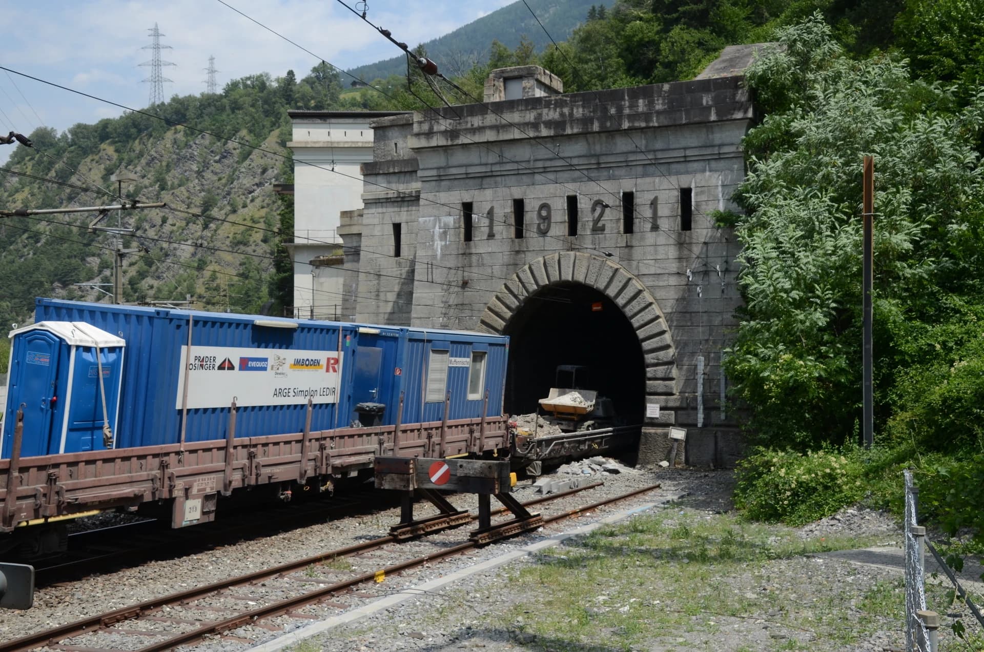 Un train de construction bleu, avec conteneurs et toilettes portables, est garé sur des voies devant l'entrée d'un tunnel ferroviaire en pierre grise, marqué des chiffres "19121", avec en arrière-plan des montagnes verdoyantes et boisées.
