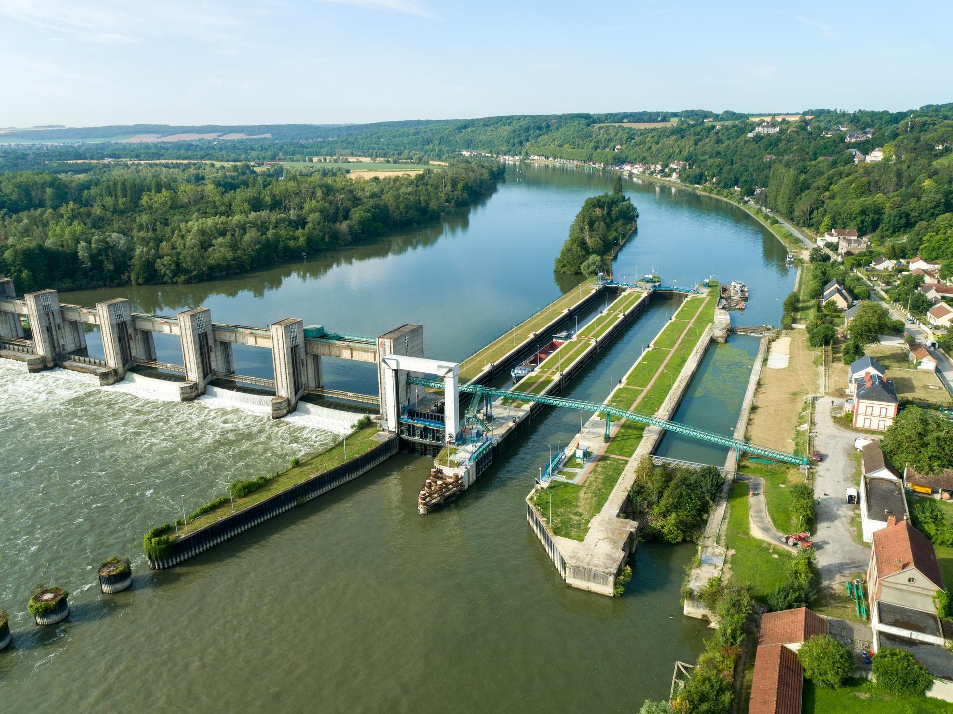 Vue aérienne d'un barrage en béton déversant de l'eau turbulente, jouxtant des écluses verdoyantes avec un pont, le tout le long d'une large rivière bordée par un village et des forêts vallonnées sous un ciel lumineux.