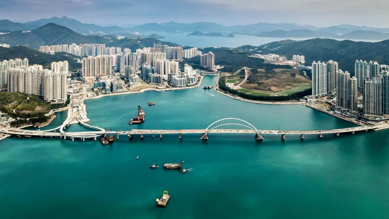 Vue aérienne d'une ville côtière dense avec des gratte-ciel, un grand pont en arc enjambant un plan d'eau et des montagnes à l'arrière-plan.