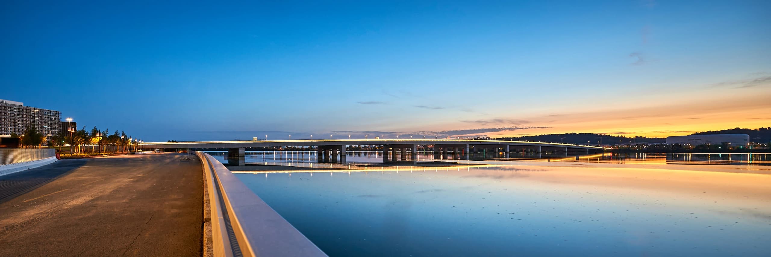 Vue panoramique d'un pont moderne illuminé s'étendant sur des eaux calmes au crépuscule, sous un ciel dégradé du bleu profond à l'orange et au jaune. Les lumières du pont et les bâtiments lointains, dont une arène, se reflètent sur la surface de l'eau, avec une promenade pavée en premier plan.