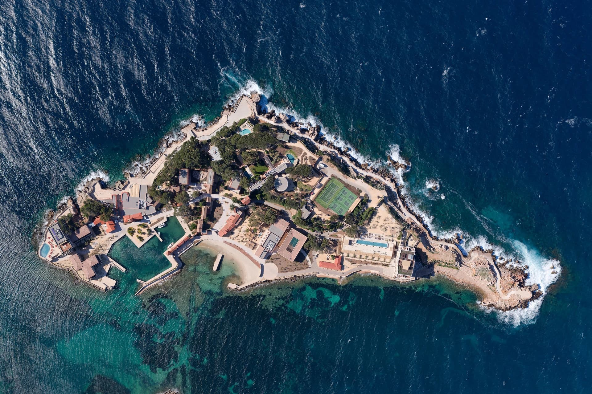Vue aérienne d'un complexe hôtelier de luxe sur une île avec court de tennis et port, entourée d'une mer bleue.