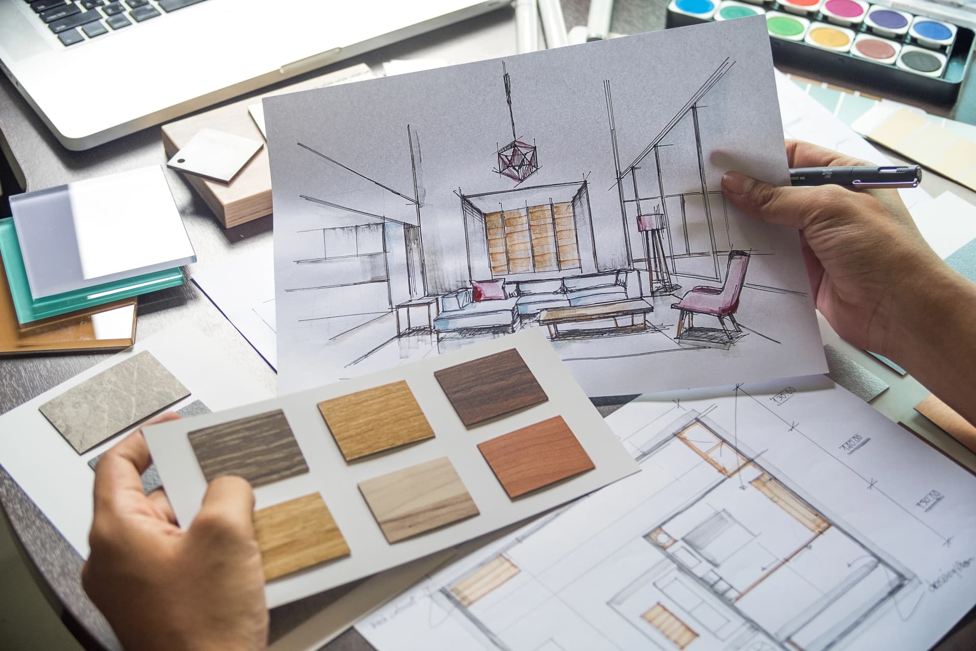 Des mains tiennent un croquis détaillé d'un salon moderne et une planche d'échantillons de finitions en bois, sur un bureau encombré de plans architecturaux, d'un ordinateur portable et d'une palette de peintures.