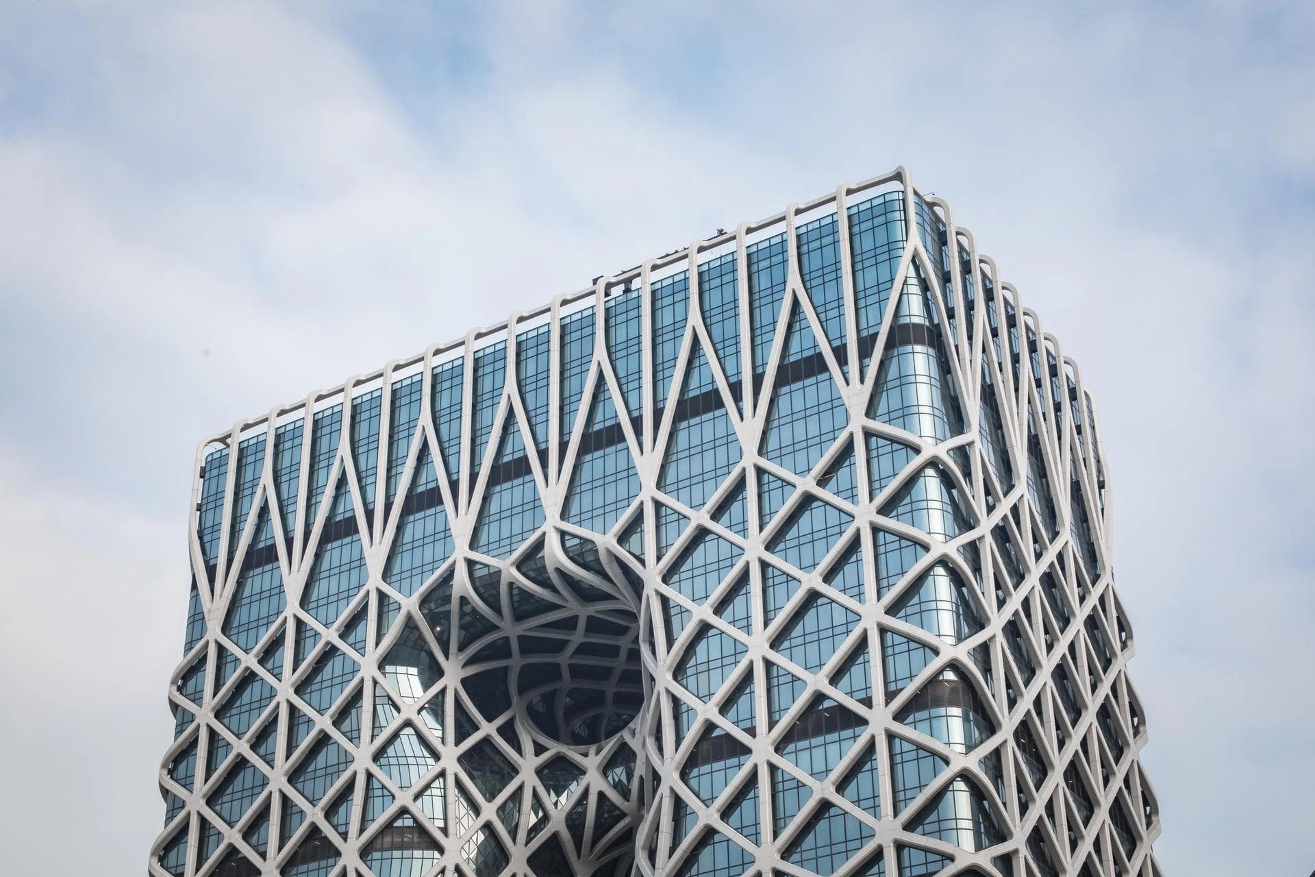 Un immeuble moderne à l'architecture complexe, doté d'une façade en verre bleu recouverte d'une structure externe blanche formant un motif géométrique en treillis, avec une grande ouverture incurvée visible sur un côté.