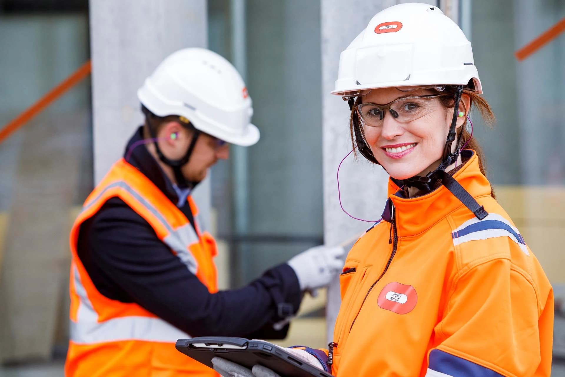 Femme souriante portant un casque, des lunettes de sécurité et un gilet orange sur un chantier, tenant une tablette, avec un collègue en arrière-plan.
