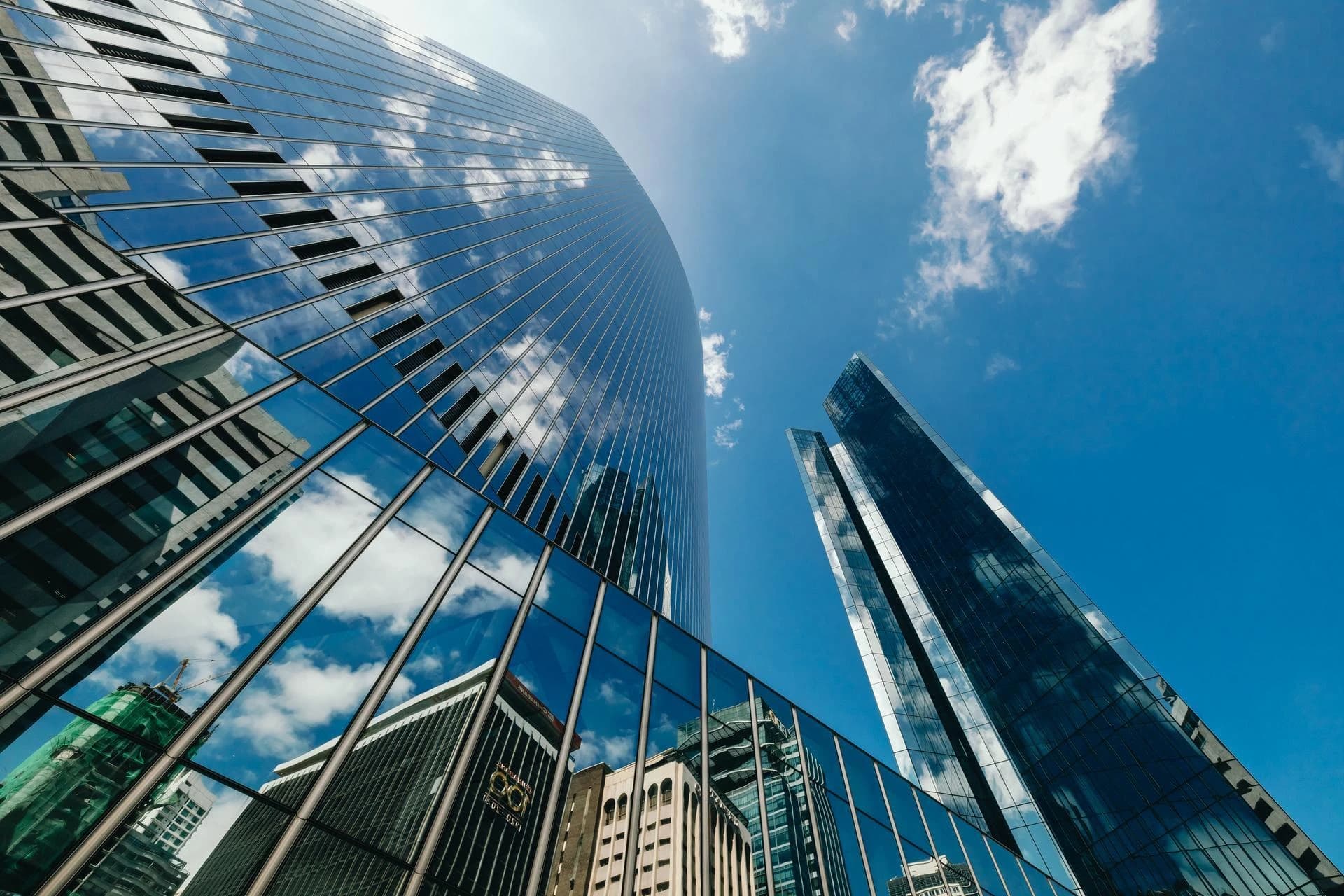 Trois gratte-ciel modernes aux façades de verre bleues s'élèvent vers un ciel d'un bleu éclatant parsemé de nuages blancs, qui se reflètent vivement sur les bâtiments. La photo est prise d'en bas, soulignant l'architecture contemporaine et la luminosité ambiante.
