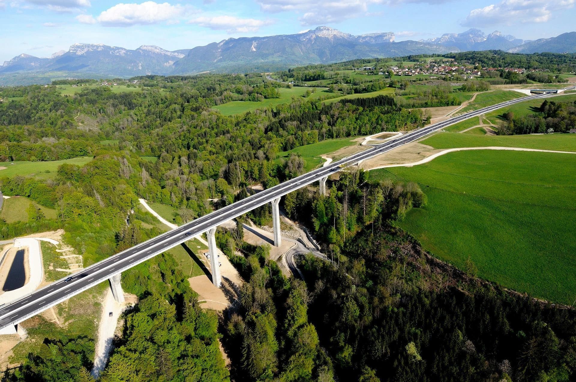 Vue aérienne d'un long viaduc autoroutier gris s'étendant à travers une vaste étendue de forêts et de prairies vertes, avec des montagnes enneigées et un village en arrière-plan sous un ciel bleu nuageux.