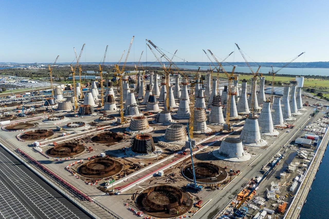 Vue aérienne d'un vaste chantier de construction montrant des dizaines de structures coniques en béton gris en cours d'édification, entourées de nombreuses grues à tour jaunes sous un ciel bleu clair, avec un plan d'eau et des terres verdoyantes en arrière-plan.