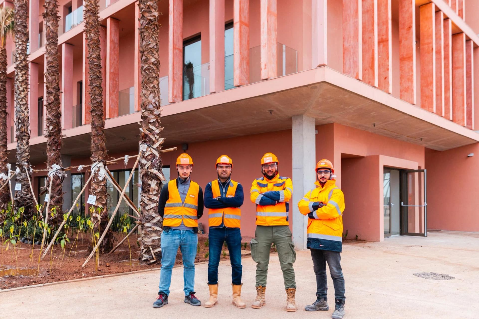 Quatre hommes vêtus de casques et de gilets orange de haute visibilité posent devant un bâtiment moderne couleur terre cuite avec des palmiers et jeunes pousses sur un chantier.