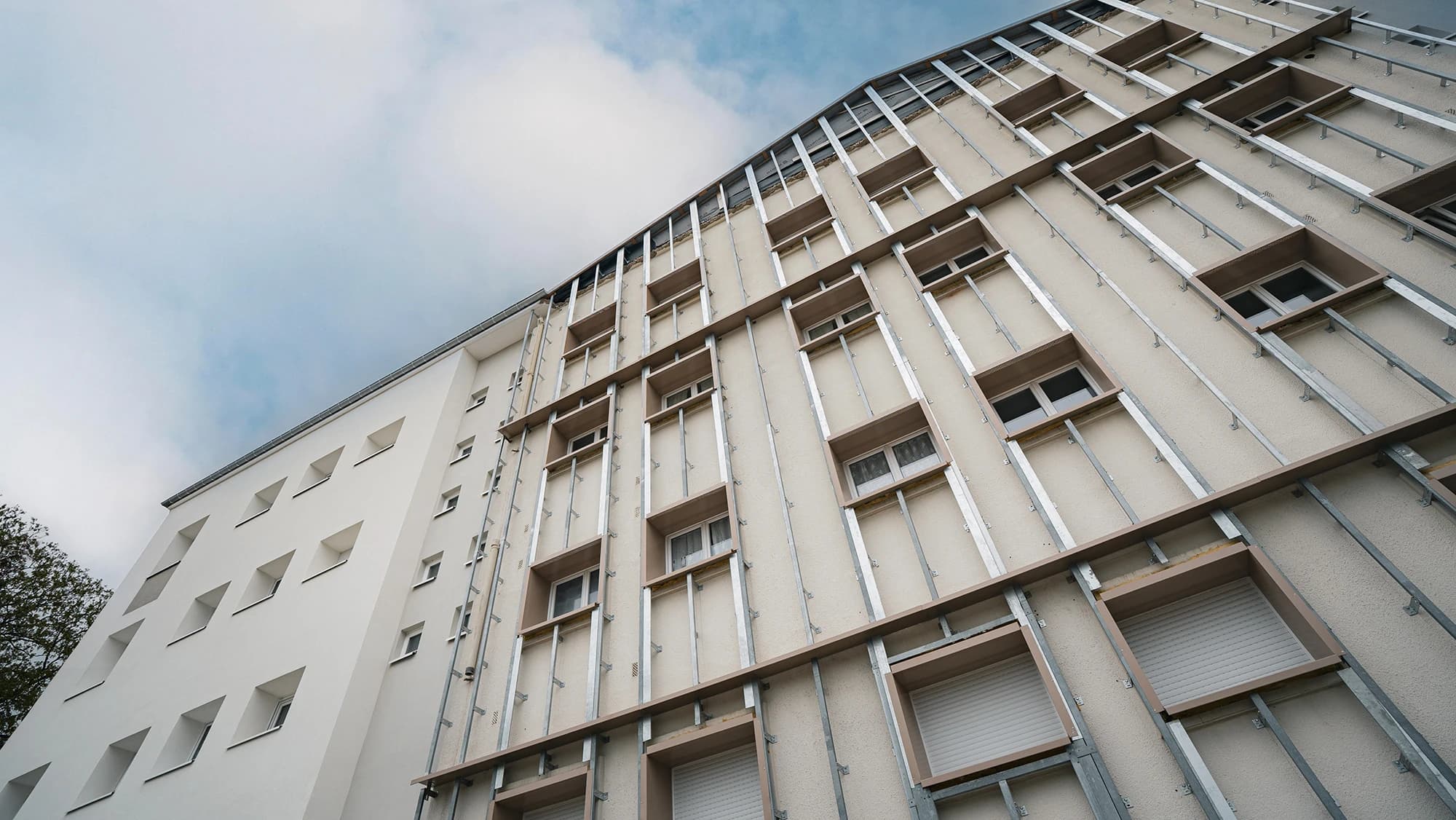 Façade d'un bâtiment en cours de rénovation, montrant une section blanche moderne à gauche et une section de droite dont le mur beige est recouvert d'une armature métallique pour une nouvelle isolation ou un nouveau revêtement, sous un ciel nuageux.