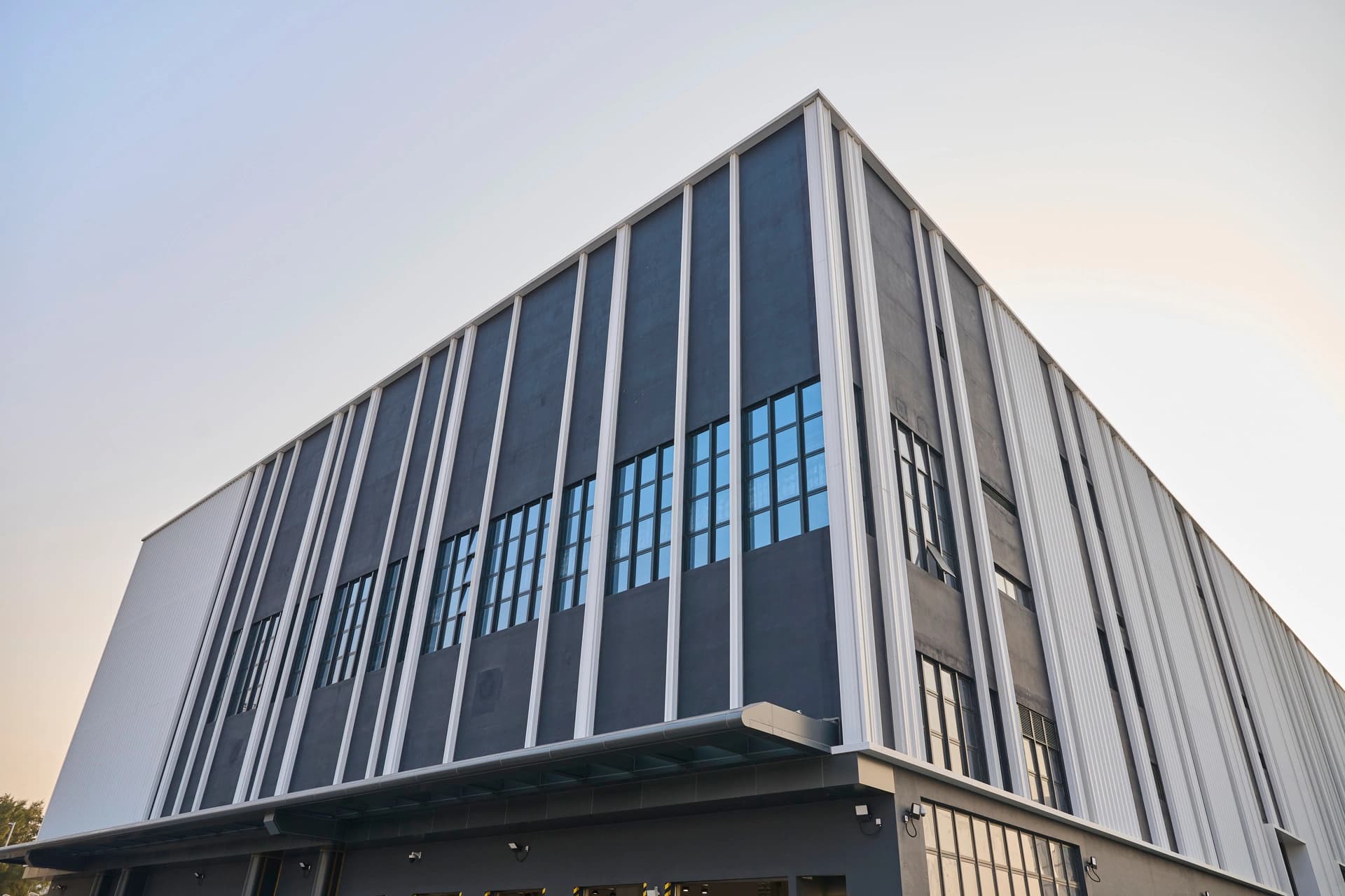Vue en contre-plongée d'un bâtiment industriel moderne gris anthracite et blanc, caractérisé par des bandes verticales et des fenêtres bleues réfléchissantes sous un ciel lumineux.