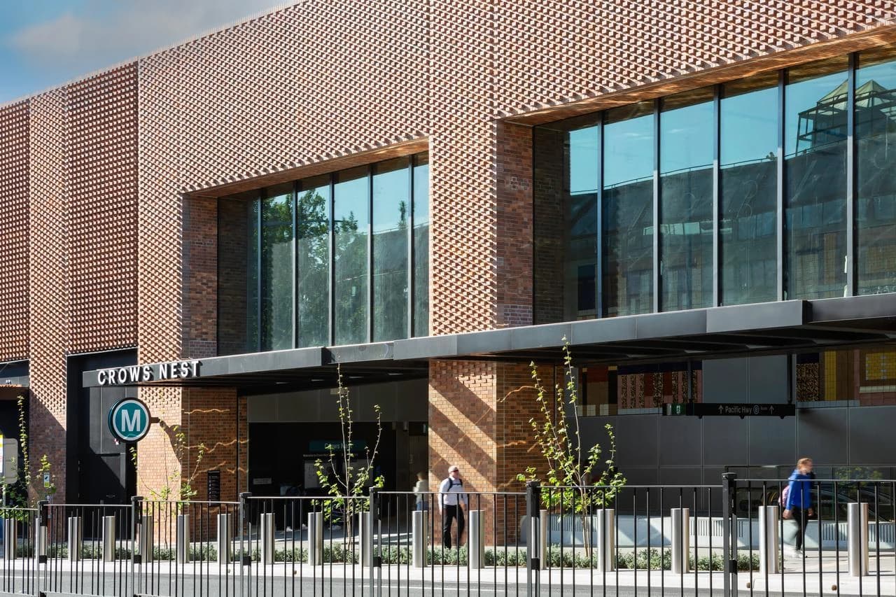 Façade moderne de la station de métro Crows Nest avec des briques texturées, de grandes fenêtres et une enseigne de métro, avec des passants.