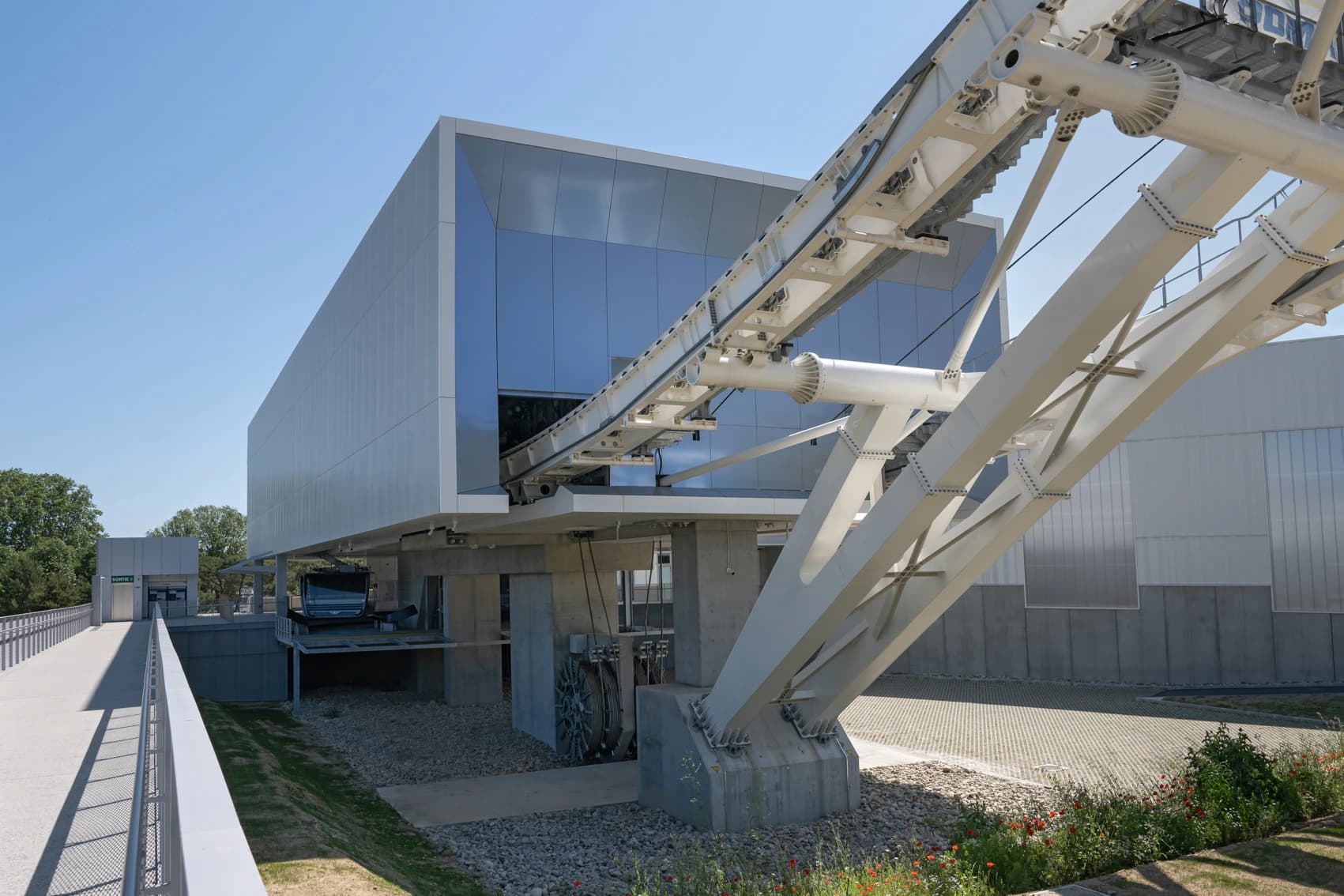 A modern building with reflective blue glass and light grey panels features a white automated people mover track curving into its entrance. A pedestrian walkway leads to a small exit building on the left, all under a clear blue sky.