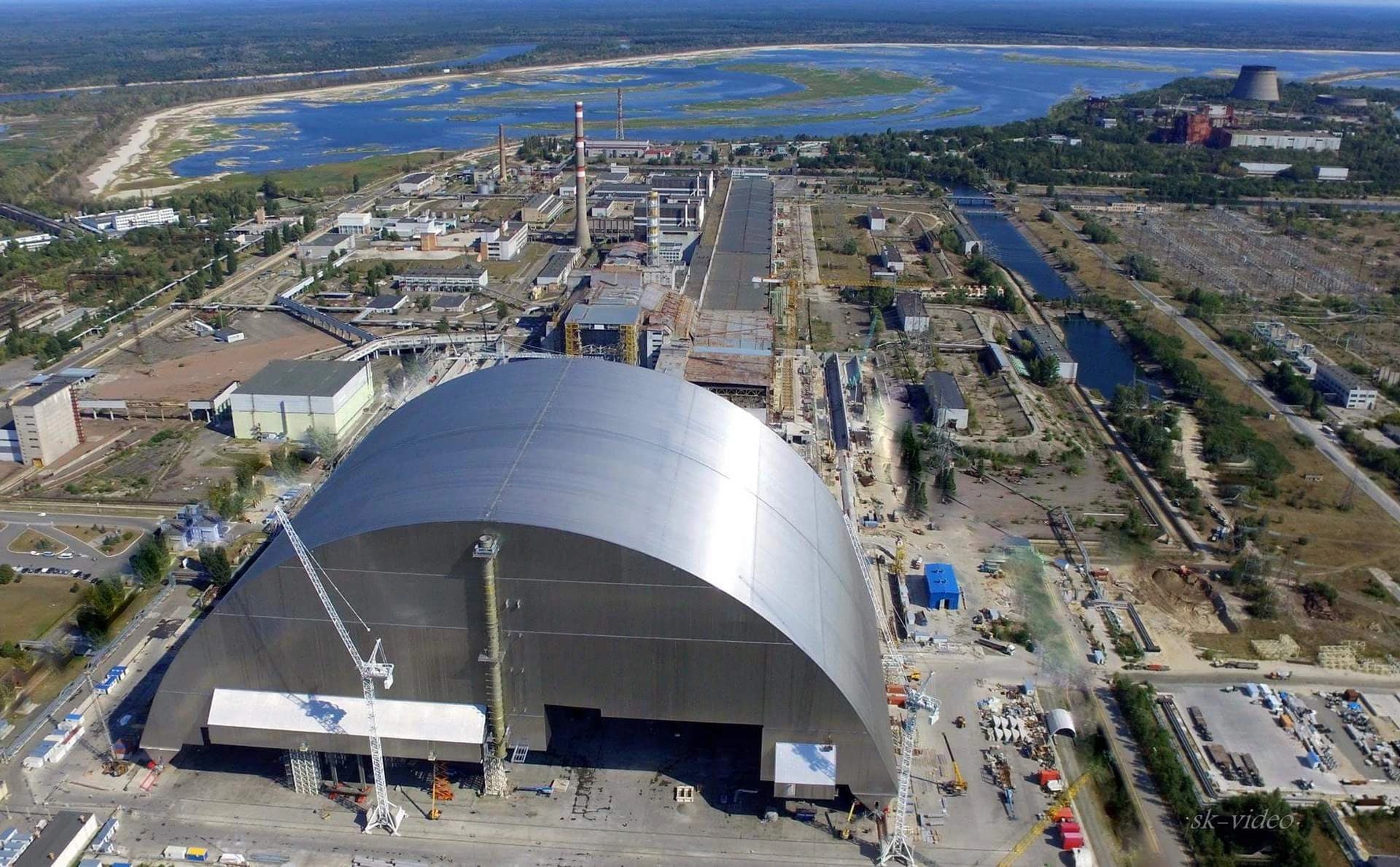 Vue aérienne du sarcophage en forme d'arche, gris métallisé, du réacteur de Tchernobyl, entouré d'autres bâtiments industriels, de zones vertes et du fleuve Pripyat sous un ciel bleu.