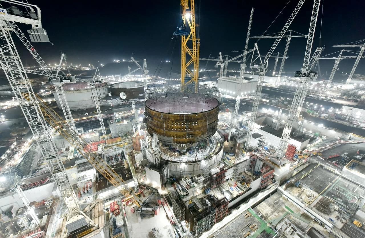Vue aérienne nocturne d'un vaste chantier de construction, où une grue jaune géante soulève une imposante structure cylindrique métallique et dorée, entourée de nombreux bâtiments inachevés et autres grues éclairés par des lumières vives.