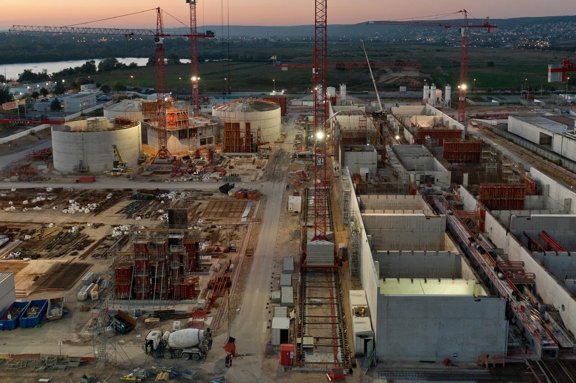 Vue aérienne d'un immense chantier de construction industriel au crépuscule, montrant de nombreuses grues rouges au-dessus de structures en béton massives, dont des bâtiments circulaires, sous un ciel orangé avec des lumières artificielles.