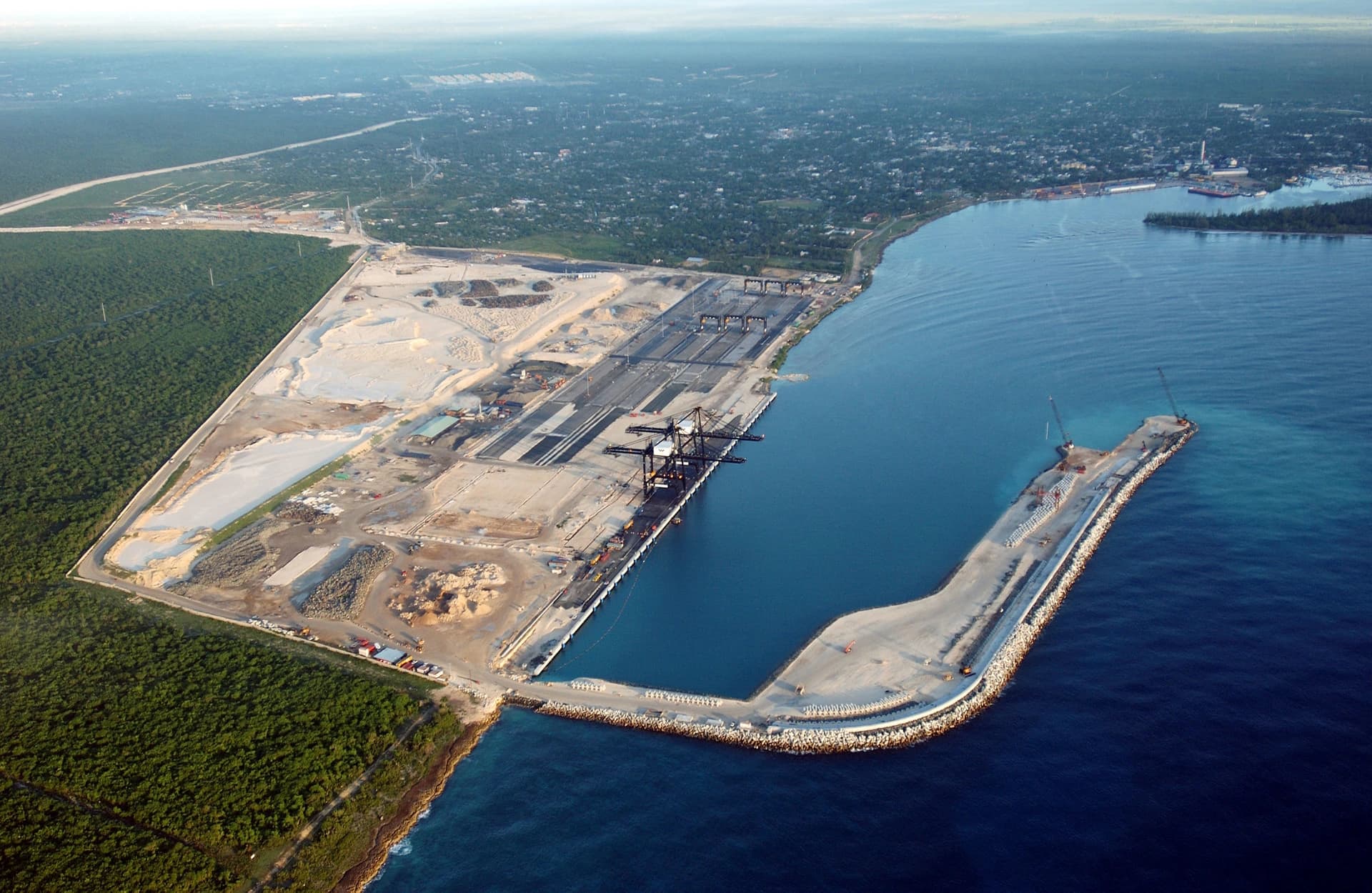 Vue aérienne d'un grand port de conteneurs avec plusieurs grues noires alignées le long des quais, adjacent à un vaste terrain en construction clair et une dense forêt verte. Un long brise-lames incurvé s'avance dans les eaux bleues de l'océan, protégeant l'entrée du port.