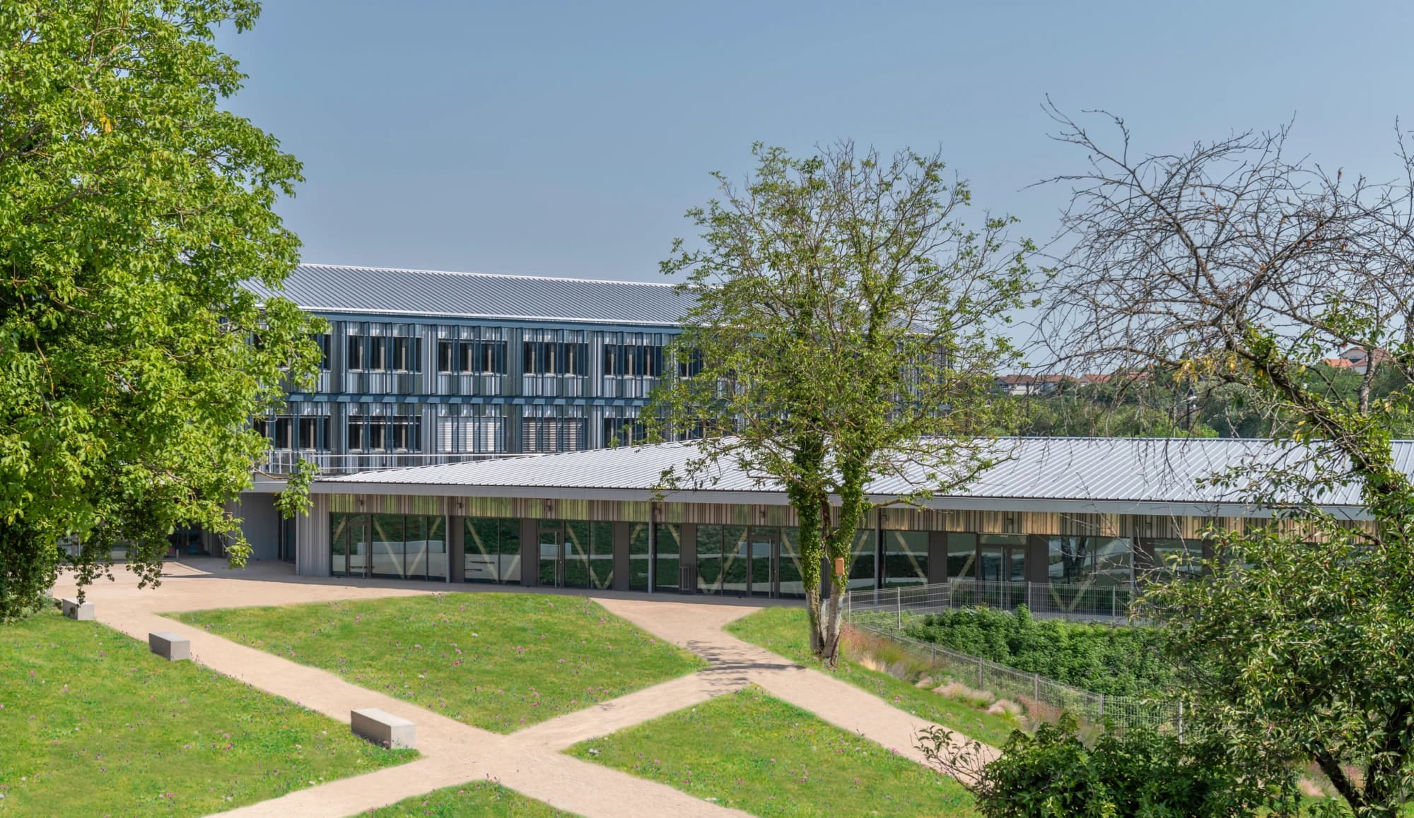 Vue extérieure d'un campus moderne avec un grand bâtiment bleu-gris et un pavillon en verre, entourés de pelouses vertes, de chemins de gravier et d'arbres sous un ciel clair.