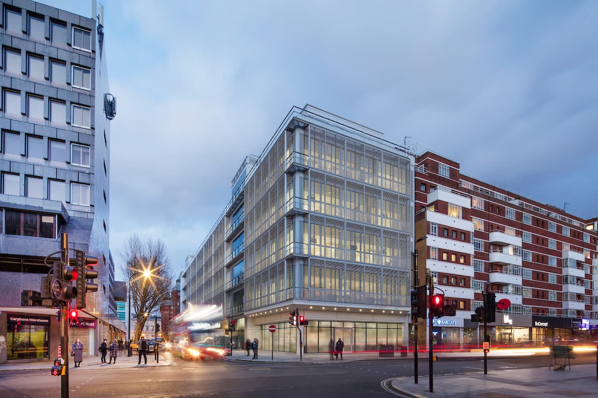 Un bâtiment moderne à persiennes gris clair et illuminé se dresse sur un coin de rue urbain au crépuscule, avec un trafic flou générant des traînées lumineuses rouges et des piétons, entouré d'immeubles variés.