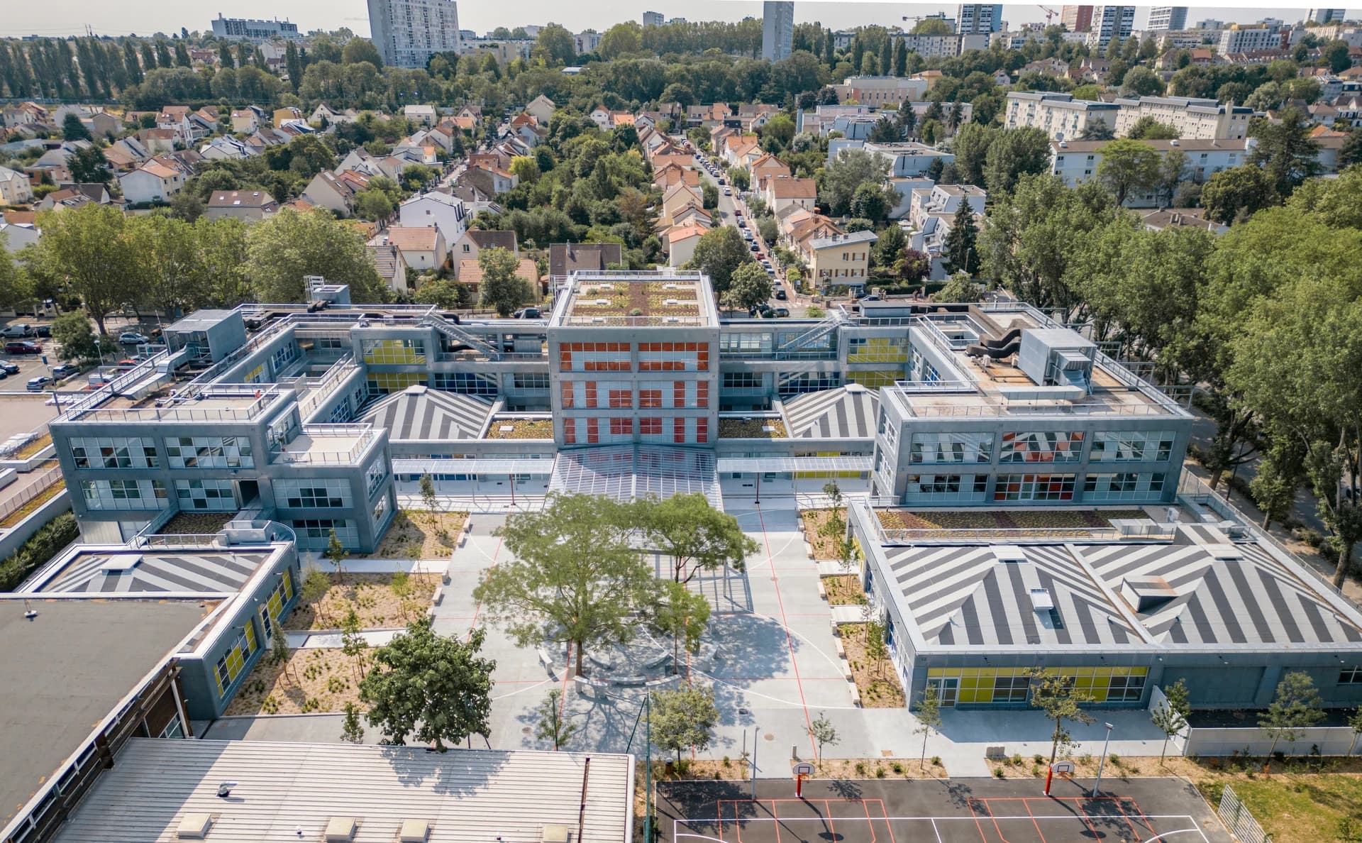 Vue aérienne d'un grand complexe scolaire moderne gris avec des accents jaunes et rouges, des toits verts et des toits rayés, centré autour d'une cour pavée arborée, entouré de maisons résidentielles et d'arbres sous un ciel clair.