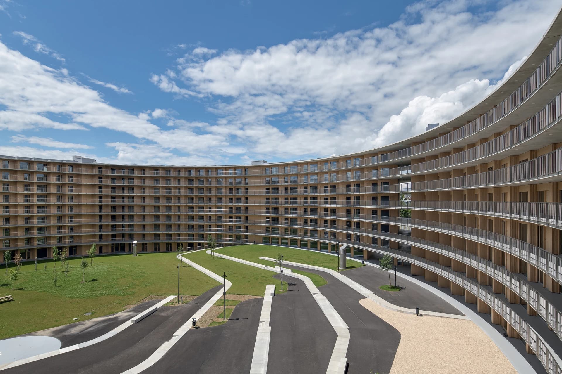 Grand bâtiment moderne incurvé, de couleur bois clair avec balcons et fenêtres, entourant une pelouse verte traversée par des chemins sombres, sous un ciel bleu parsemé de nuages blancs.
