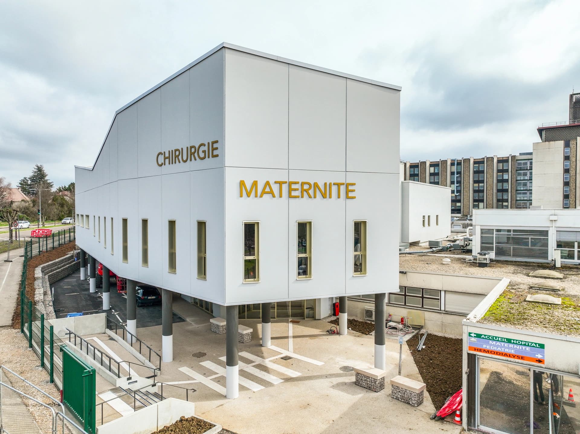 Aile hospitalière moderne avec enseignes Chirurgie et Maternité, sur pilotis blancs.