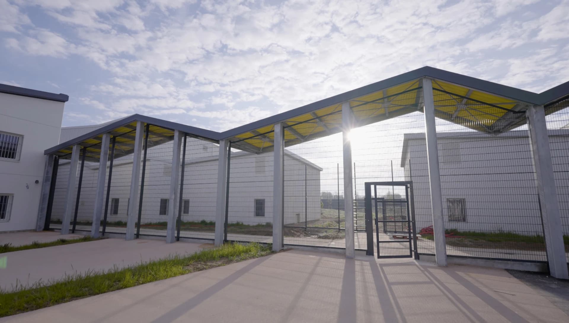 Vue extérieure d'un complexe moderne aux murs blancs et hautes clôtures métalliques, avec un passage couvert soutenu par des piliers gris et un plafond jaune vif, sous un ciel nuageux et ensoleillé.