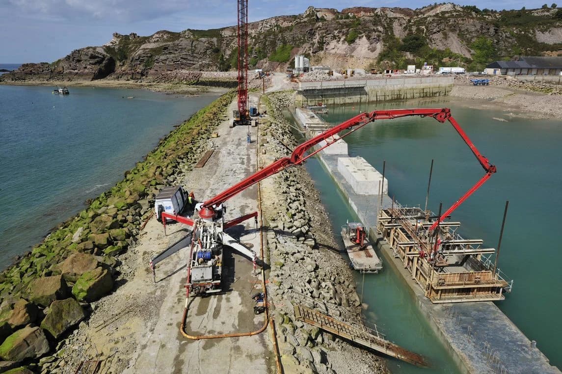 Vue aérienne d'un chantier de construction maritime où une pompe à béton rouge sur une digue rocheuse déverse du ciment dans des structures en coffrage flottant sur une eau turquoise. À l'arrière-plan, on aperçoit une grue, des falaises et une carrière.