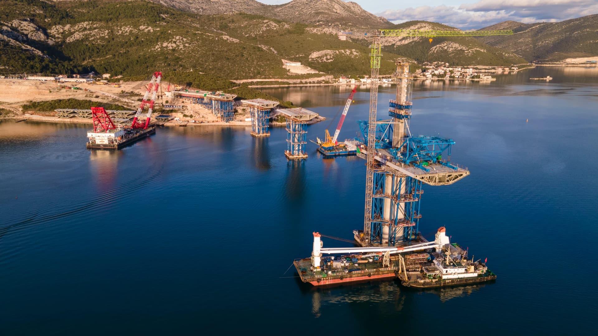 Vue aérienne d’un chantier de construction d’un pont maritime. Plusieurs piles en béton sont en cours d’installation en mer à l’aide de grues flottantes et d’équipements lourds. Le site se situe dans une zone côtière montagneuse avec une eau calme, illustrant un projet d’ingénierie civile et de travaux publics de grande envergure.