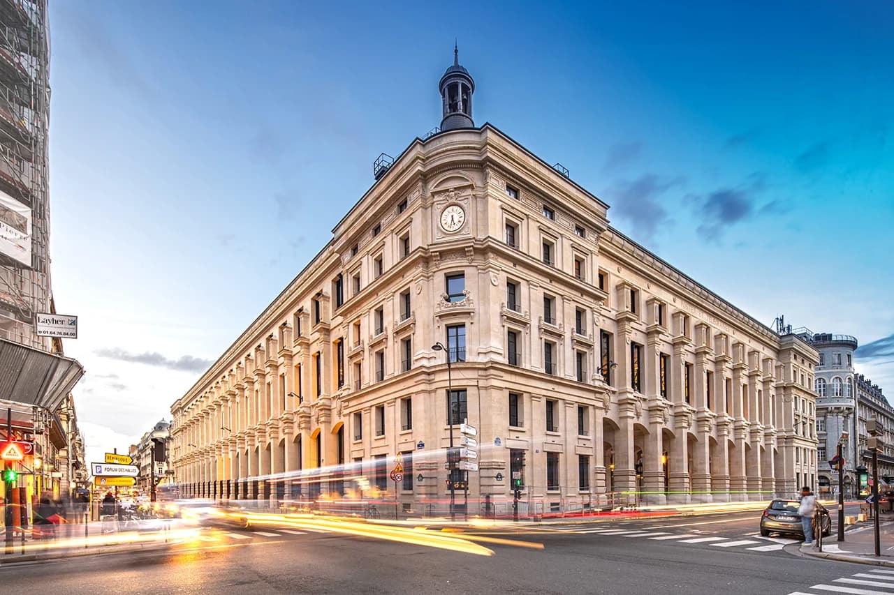 Grand bâtiment parisien à l'architecture classique en pierre claire, avec une horloge et une coupole à l'angle, au crépuscule. Des traînées lumineuses de véhicules animent un carrefour urbain, sous un ciel dégradé du bleu clair au bleu profond.