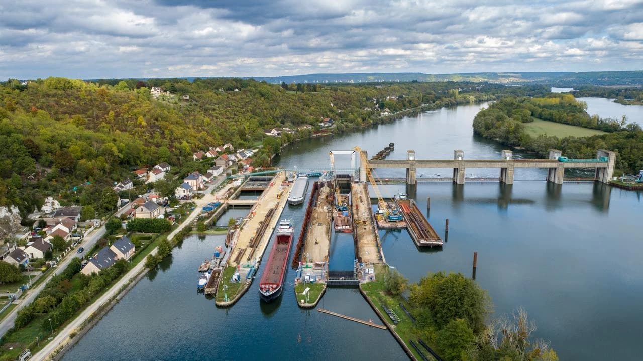 Vue aérienne d'un vaste complexe d'écluses fluviales avec des barges et grues en activité sur une large rivière, à côté d'un barrage en béton et d'un village. Le paysage est entouré de collines verdoyantes sous un ciel partiellement nuageux.