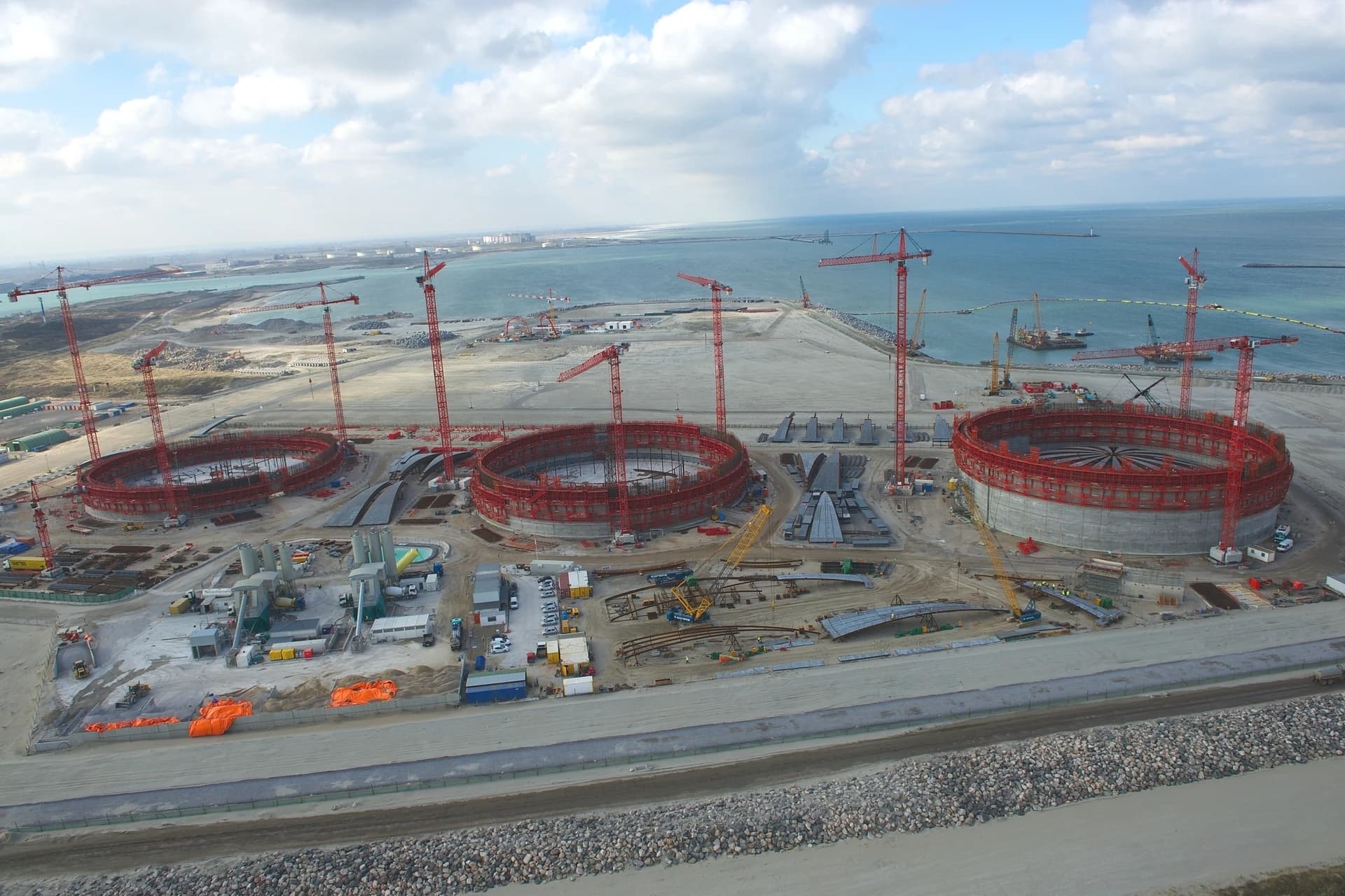 Vue aérienne d'un vaste chantier de construction près de la mer, dominé par trois grandes structures circulaires en béton avec échafaudages rouges et de nombreuses grues rouges. Le site, principalement composé de terre dégagée, s'étend jusqu'à l'océan bleu clair sous un ciel nuageux, avec des installations industrielles lointaines.