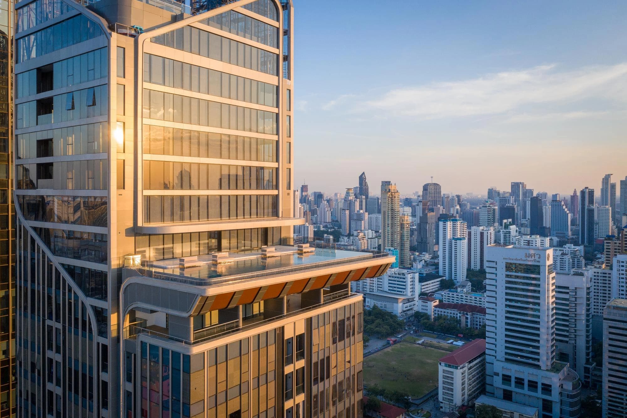 Vue aérienne d'un gratte-ciel moderne à façade vitrée, reflétant la lumière dorée du soleil sur sa piscine à débordement de toit, dominant une vaste ville aux nombreux bâtiments sous un ciel clair.