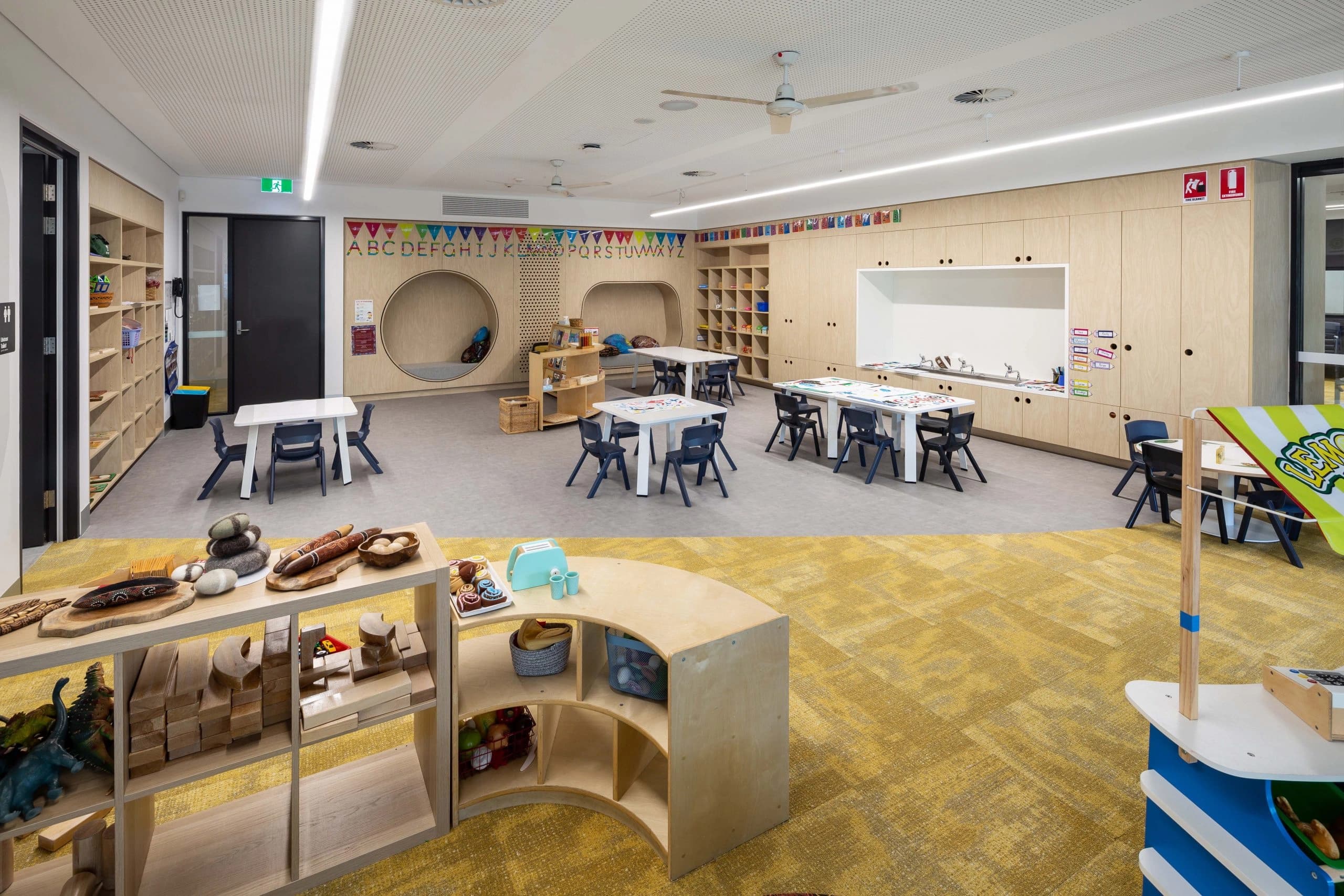 Un environnement d'apprentissage moderne et lumineux pour enfants avec des meubles en bois clair, des tables et chaises à taille d'enfant, des recoins circulaires confortables, et des étagères remplies de jouets éducatifs, complété par un sol en dalles grises et un tapis jaune.