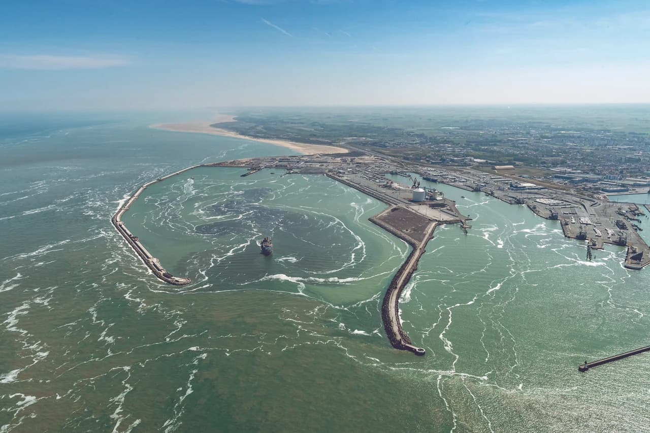 Vue aérienne d'un grand port à conteneurs où des milliers de conteneurs colorés sont empilés le long de quais gris, desservis par de massives grues noires chargeant un cargo vert dans des eaux turquoise.