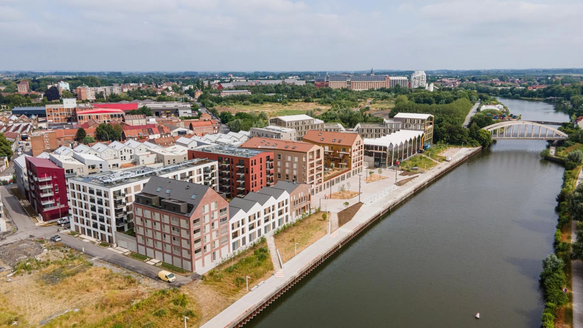 Vue aérienne d'un développement urbain moderne avec des bâtiments résidentiels à plusieurs étages de couleurs rouge, blanche et brique le long d'un canal sombre bordé d'une large promenade. Un pont en arc blanc et un paysage urbain verdoyant s'étendent en arrière-plan sous un ciel nuageux.