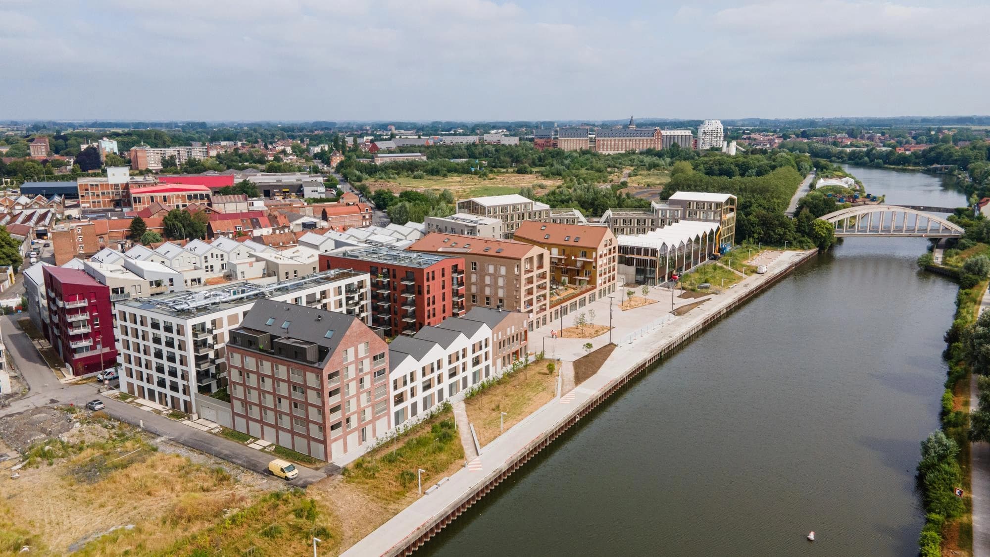 Vue aérienne d'un développement urbain moderne avec des bâtiments résidentiels à plusieurs étages de couleurs rouge, blanche et brique le long d'un canal sombre bordé d'une large promenade. Un pont en arc blanc et un paysage urbain verdoyant s'étendent en arrière-plan sous un ciel nuageux.