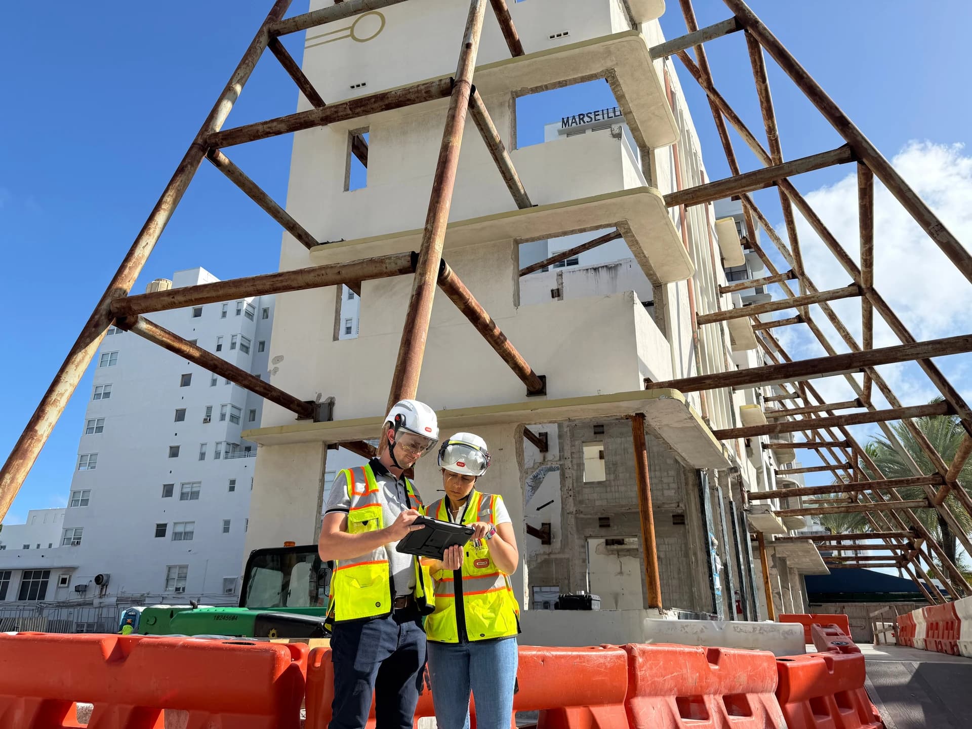 Deux travailleurs du bâtiment en casques et gilets jaunes fluo consultent une tablette sur un chantier ensoleillé, devant des barrières oranges, un bâtiment blanc en rénovation et une grande structure de support métallique rouillée sous un ciel bleu clair.