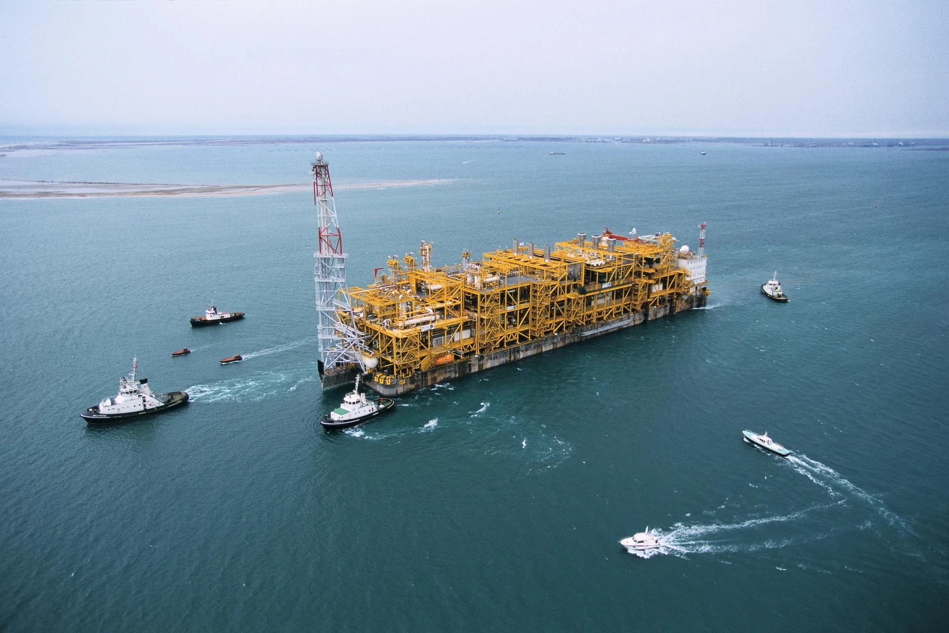 Vue aérienne d'une immense plateforme offshore jaune et blanche avec une tour rouge, remorquée par plusieurs navires dans des eaux bleu-vert sous un ciel couvert, avec des bateaux plus petits l'accompagnant.
