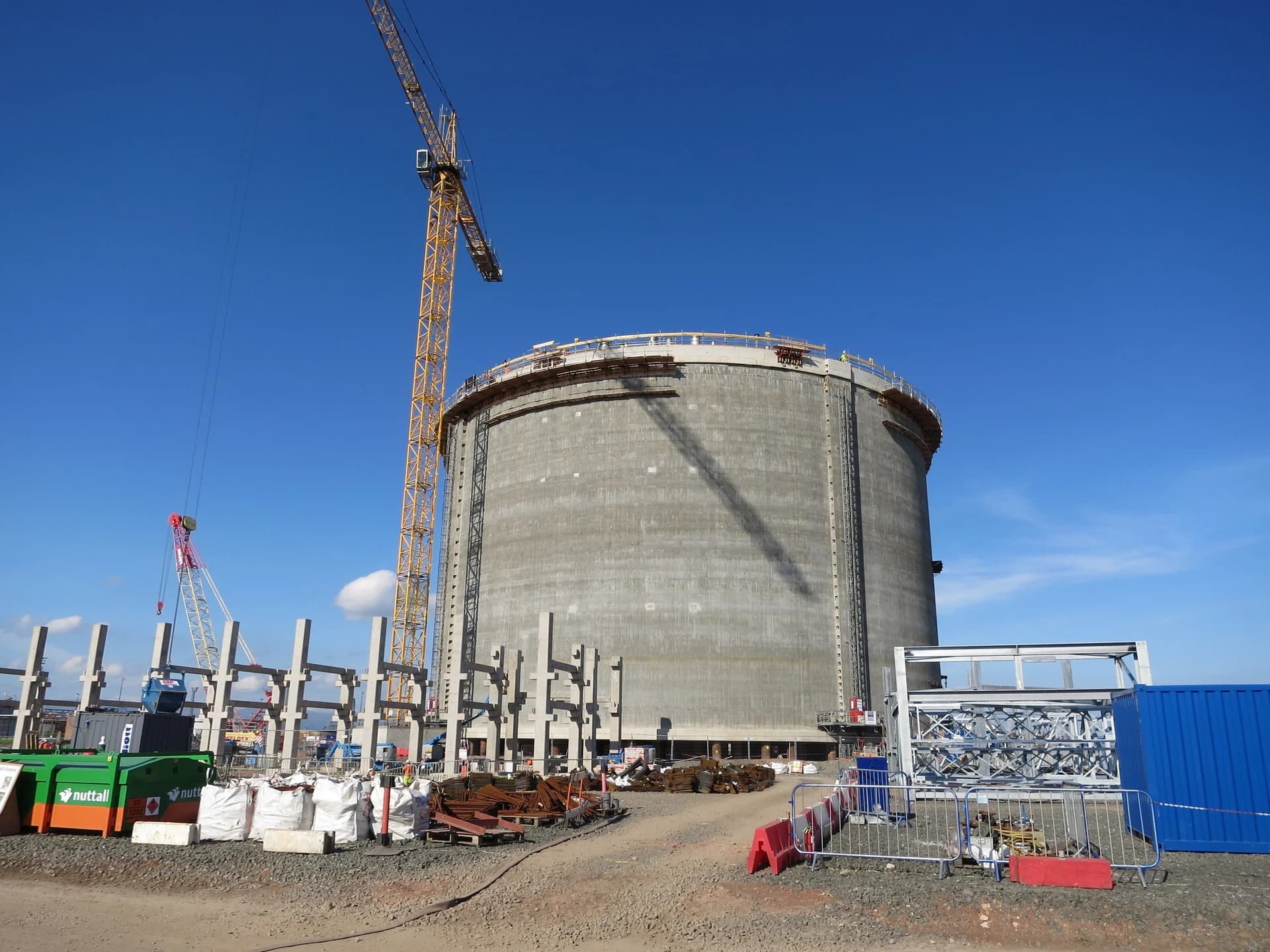 Un grand réservoir circulaire en béton gris est en construction sous un ciel bleu clair, avec une imposante grue jaune et divers matériaux de chantier au premier plan.