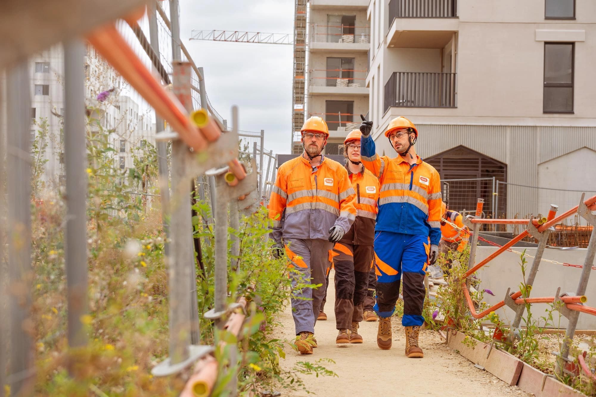 Trois ouvriers en gilets orange et bleus à haute visibilité et casques de chantier marchent sur un chemin de terre sur un site de construction, l'un d'eux pointant du doigt vers des bâtiments en construction et une grue.