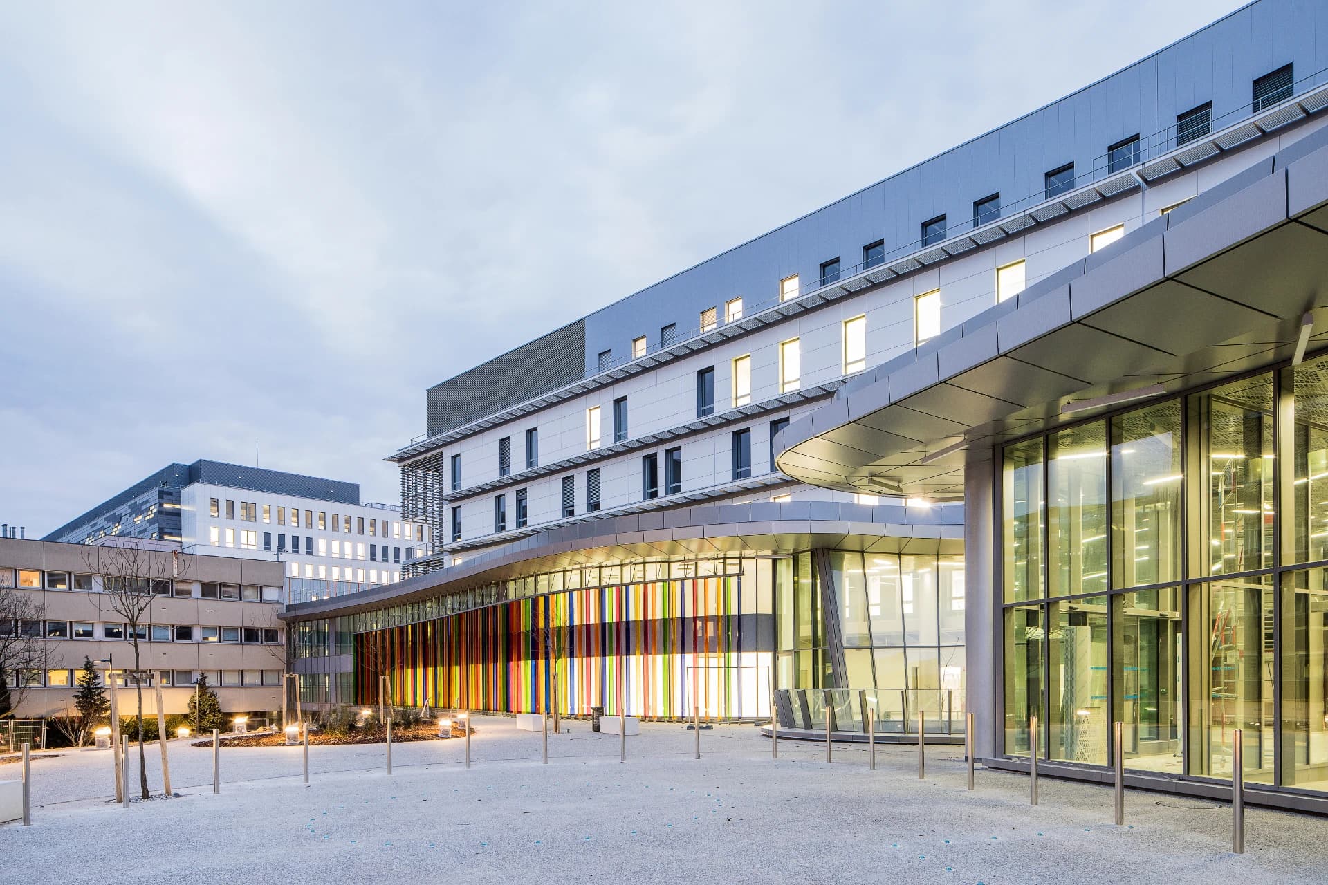 Façade moderne d'un hôpital ou d'un grand bâtiment public, avec des panneaux gris et blancs, des fenêtres éclairées et une section distinctive ornée de bandes verticales multicolores. L'édifice s'élève sous un ciel couvert, surplombant une esplanade extérieure éclairée.