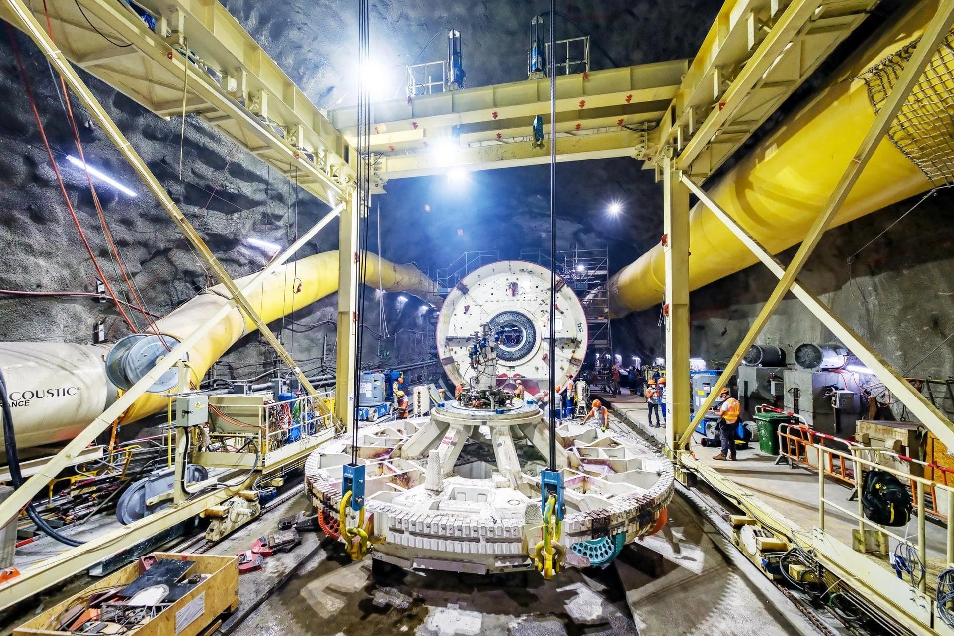 Une tête de tunnelier massive est assemblée dans un chantier souterrain, entourée de travailleurs, de structures métalliques jaunes et de conduits de ventilation lumineux.