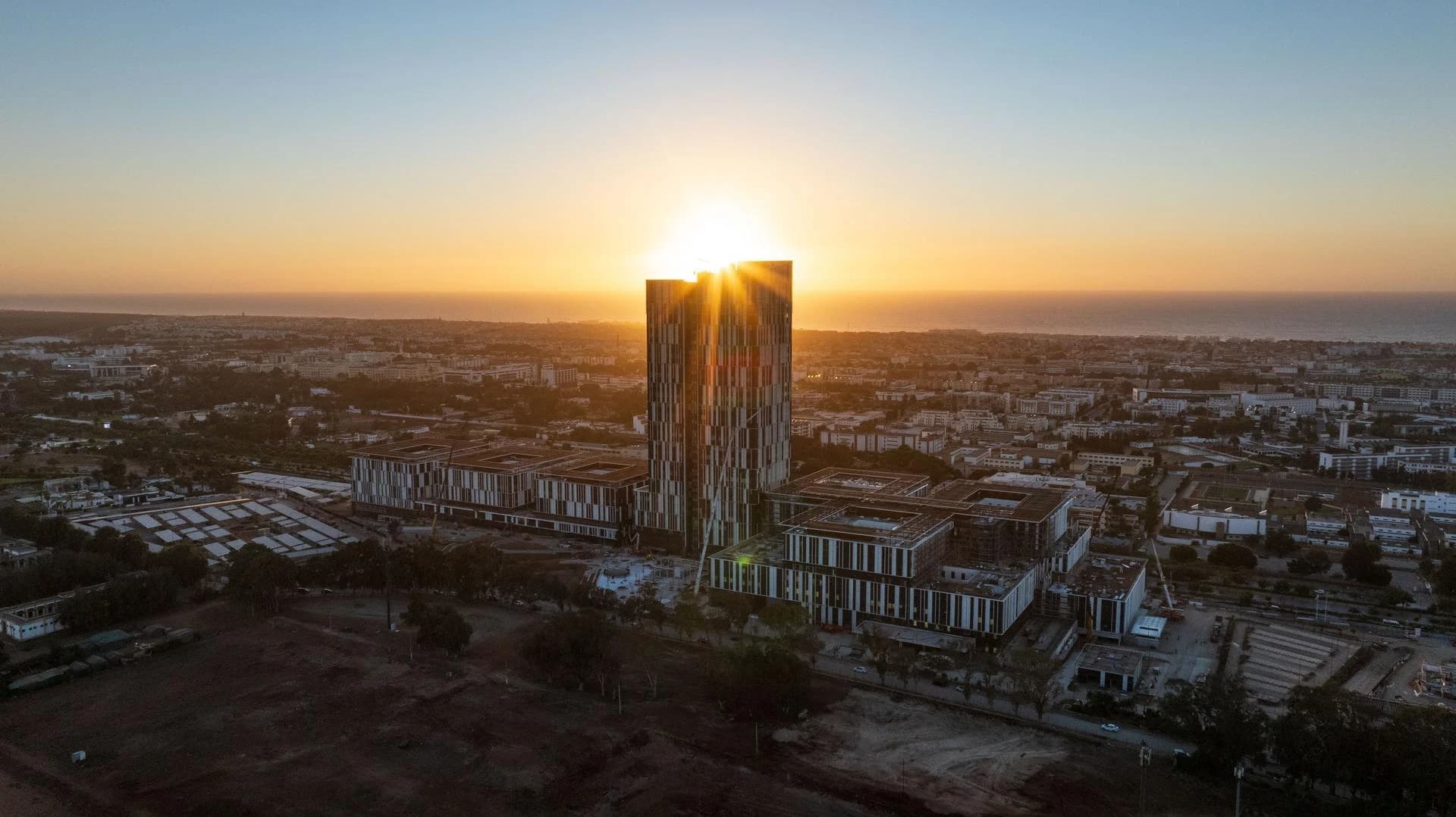 Vue aérienne d'un gratte-ciel moderne s'élevant au-dessus d'une ville au coucher du soleil, le soleil éclatant se reflétant directement derrière lui. Le paysage urbain sombre s'étend jusqu'à l'océan lointain, baigné d'une lumière orange et jaune.