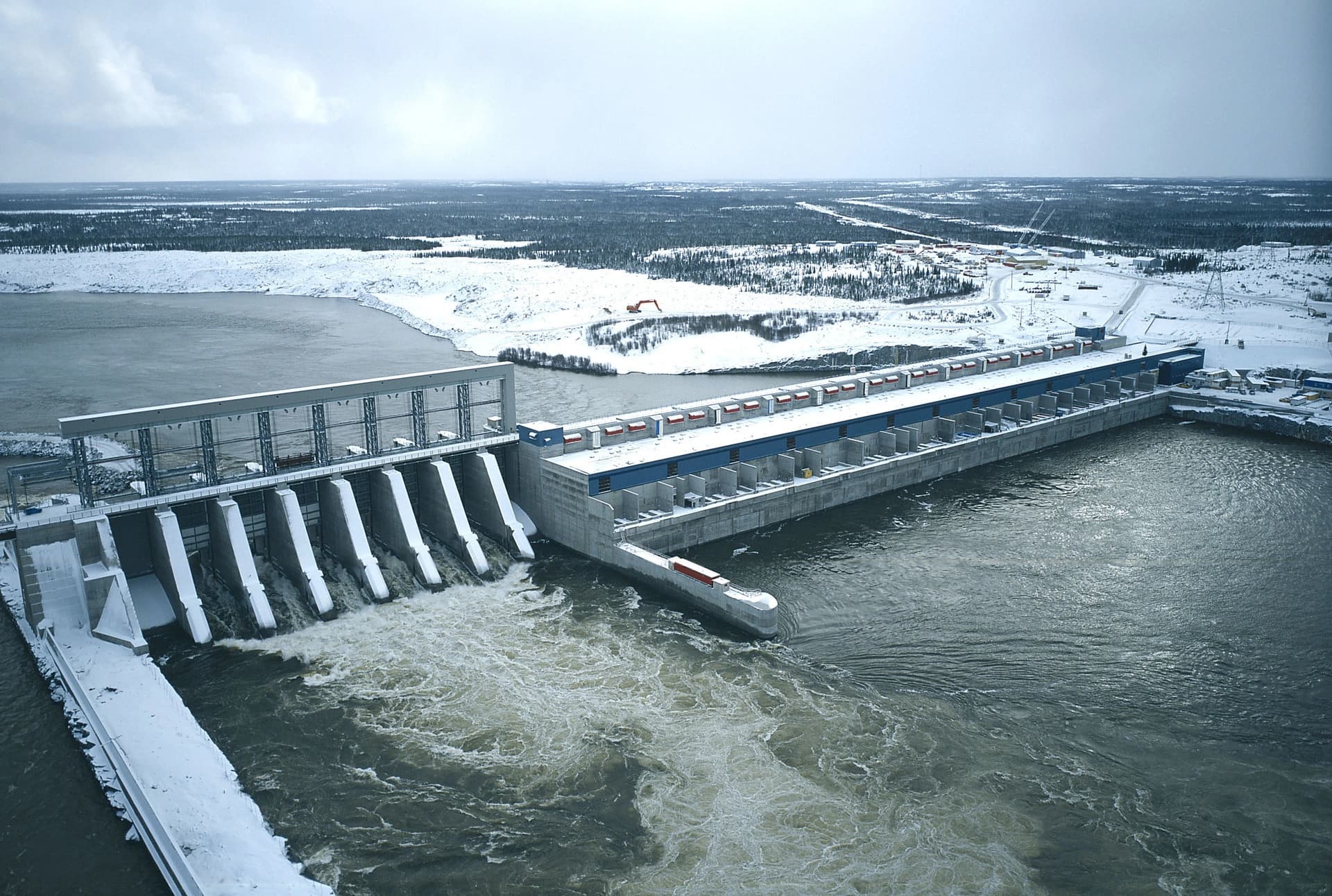 Vue aérienne d'un grand barrage hydroélectrique en hiver, avec de l'eau tumultueuse s'écoulant de son déversoir et de longues structures de centrale électrique bordant le fleuve. Le paysage environnant est couvert de neige et de vastes forêts sous un ciel nuageux.