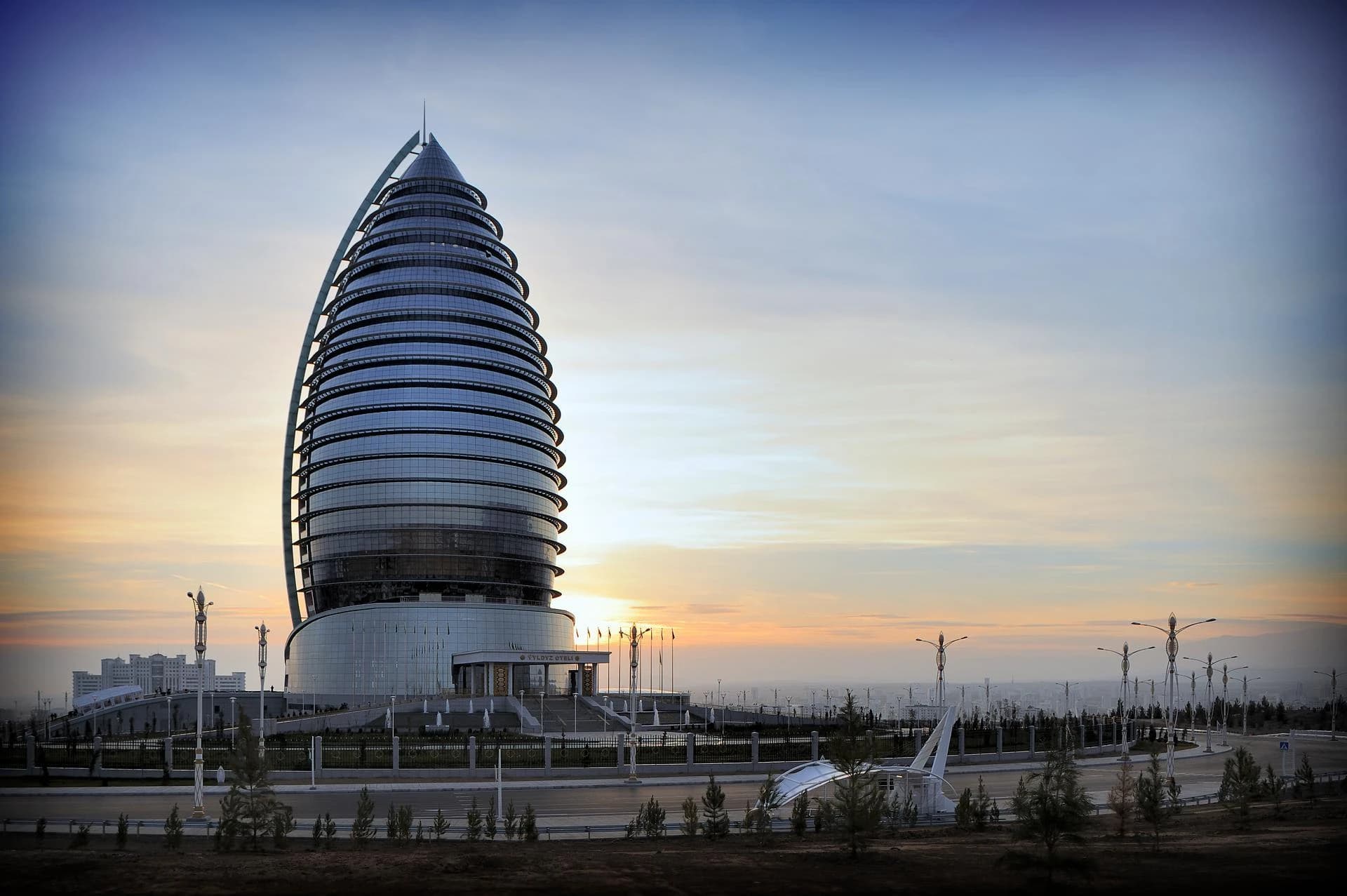 Un gratte-ciel moderne en verre, de forme conique et en spirale, domine un paysage urbain au coucher du soleil, teinté d'oranges et de bleus doux, avec des lampadaires stylisés au premier plan.
