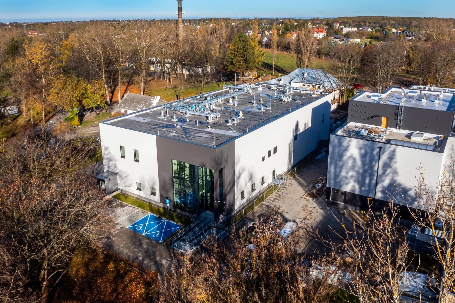 Vue aérienne d'un bâtiment moderne blanc et gris foncé avec une grande entrée vitrée, entouré d'arbres dépouillés et d'un parking avec des places handicapées bleues, sous un ciel bleu clair.