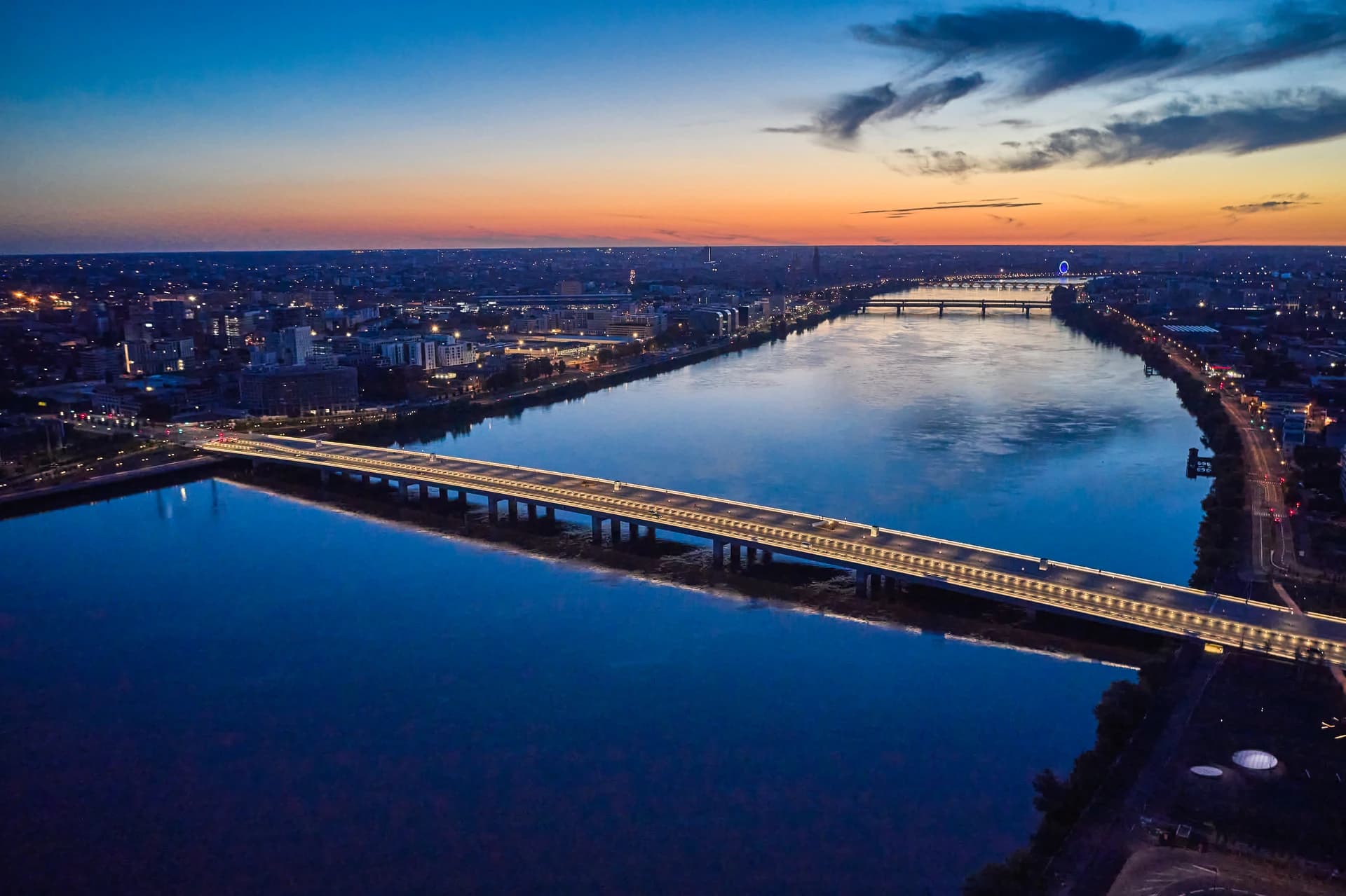 Vue aérienne d'une ville au crépuscule, avec une large rivière bleue reflétant un pont lumineux au premier plan et les lumières scintillantes de la ville sous un ciel dégradé du bleu à l'orange.