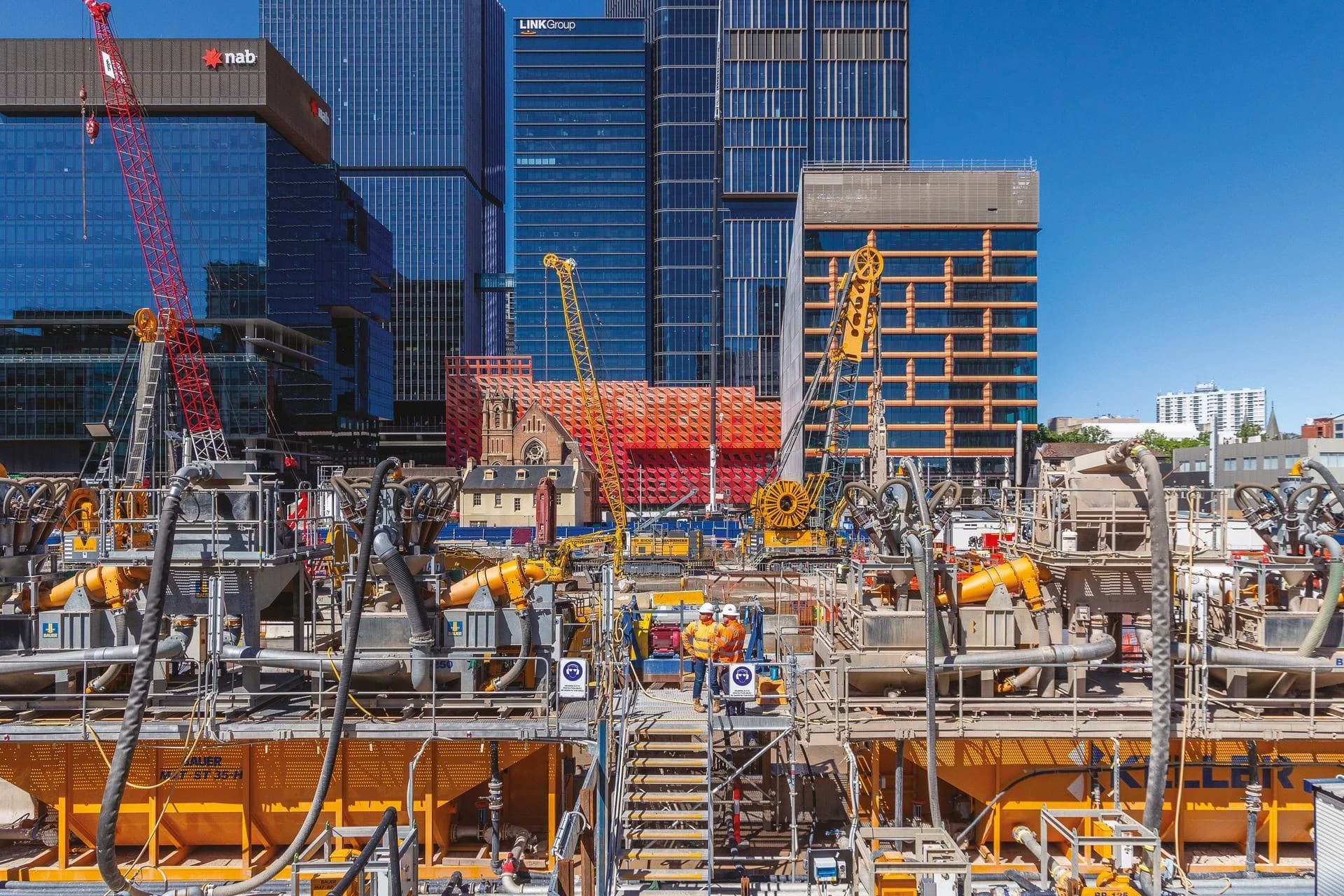 Deux ouvriers en gilets orange se tiennent sur une plateforme au milieu d'un vaste équipement de chantier jaune et gris avec des tuyaux, devant des gratte-ciel bleus modernes, une grue rouge et une église historique sous un ciel clair.